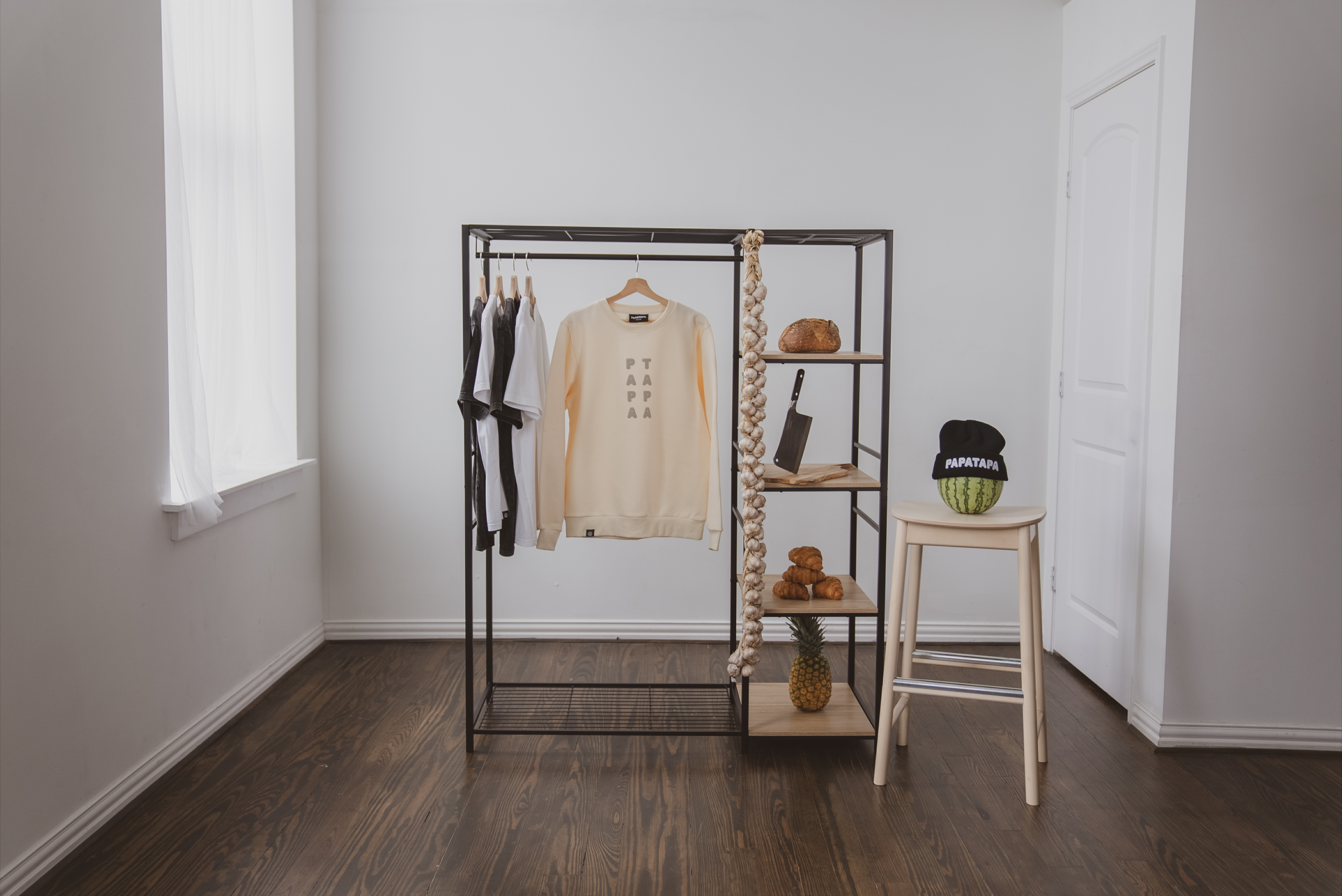 A minimalistic room with a clothing rack displaying a few t-shirts and a cream sweatshirt, a shelving unit with various items including a loaf of bread, a cleaver, and pineapples, and a small table with a watermelon wearing a black beanie that says "PAPATAPA".