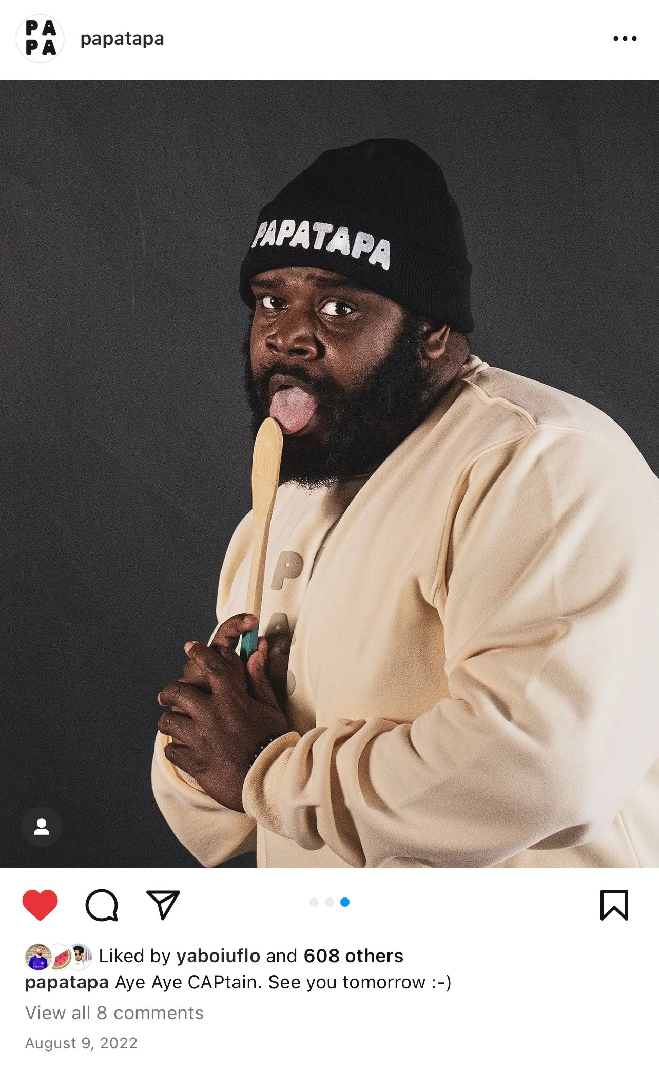 A man with a beard and mustache wearing a black beanie with 'PAPATAPA' written on it, a beige jacket, holding a wooden spoon, sticking out his tongue, and posing against a dark background.