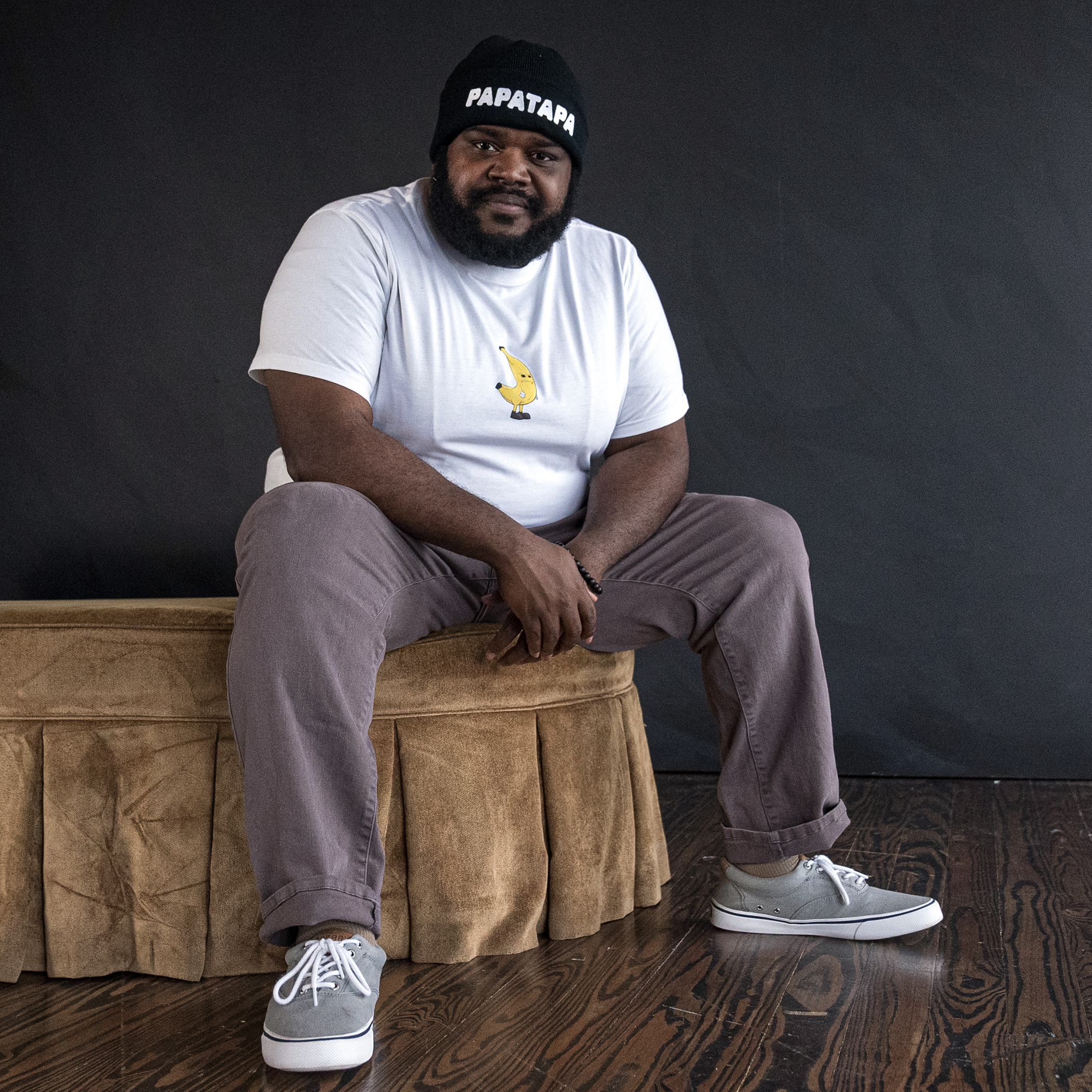 A man with a beard, sitting on a brown cushioned bench against a black background. He is wearing a black beanie with 'PAPATAPA' written on it, a white t-shirt with a banana graphic, gray pants, and gray sneakers.