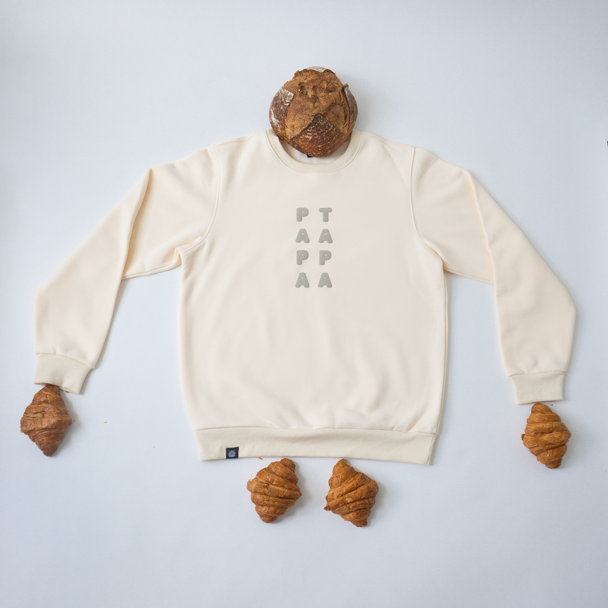 A beige sweatshirt with the word 'PAPAA' written vertically in gray letters on the front, surrounded by four croissants placed around it, with one croissant on top of the sweatshirt and three at the bottom, resembling a playful arrangement.