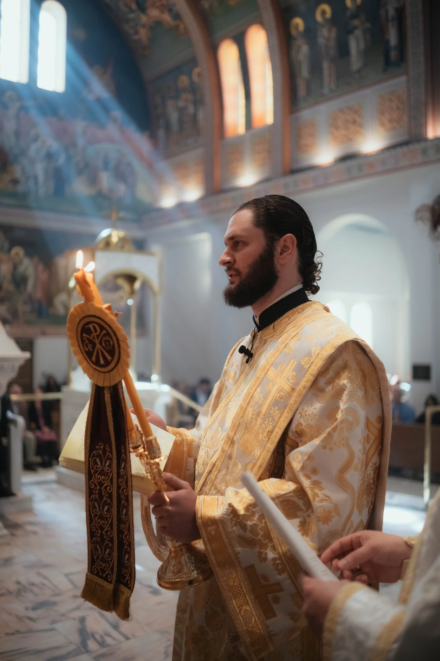 Orthodox priest in gold vestments holding a candle with a religious symbol during a church service inside a decorated church with stained glass windows and religious artwork.