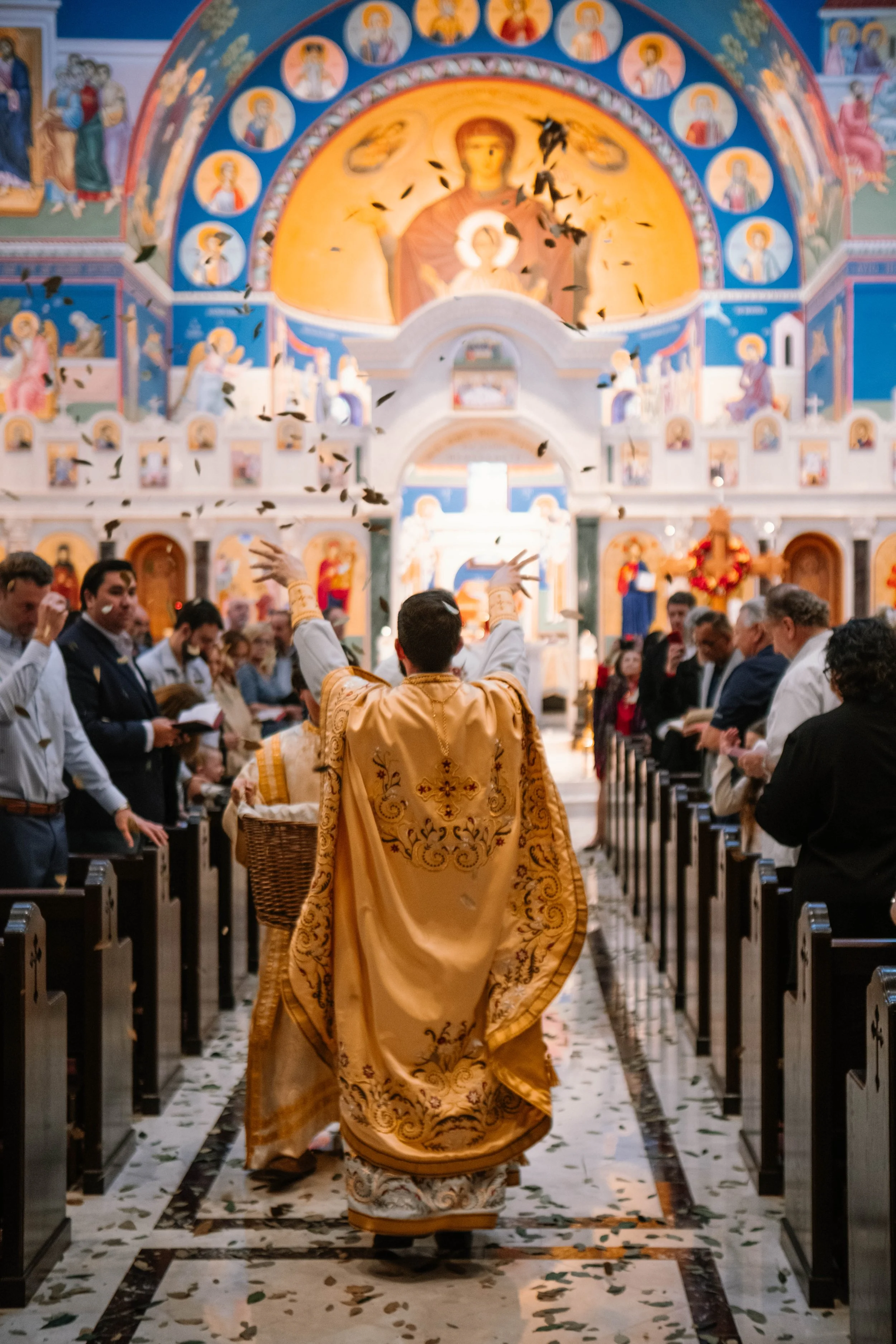 A priest in golden religious vestments leads a church service inside a colorful church with painted icons and religious murals on the walls and ceiling. Congregation members are seated in pews on either side of the aisle, some with open books, and le