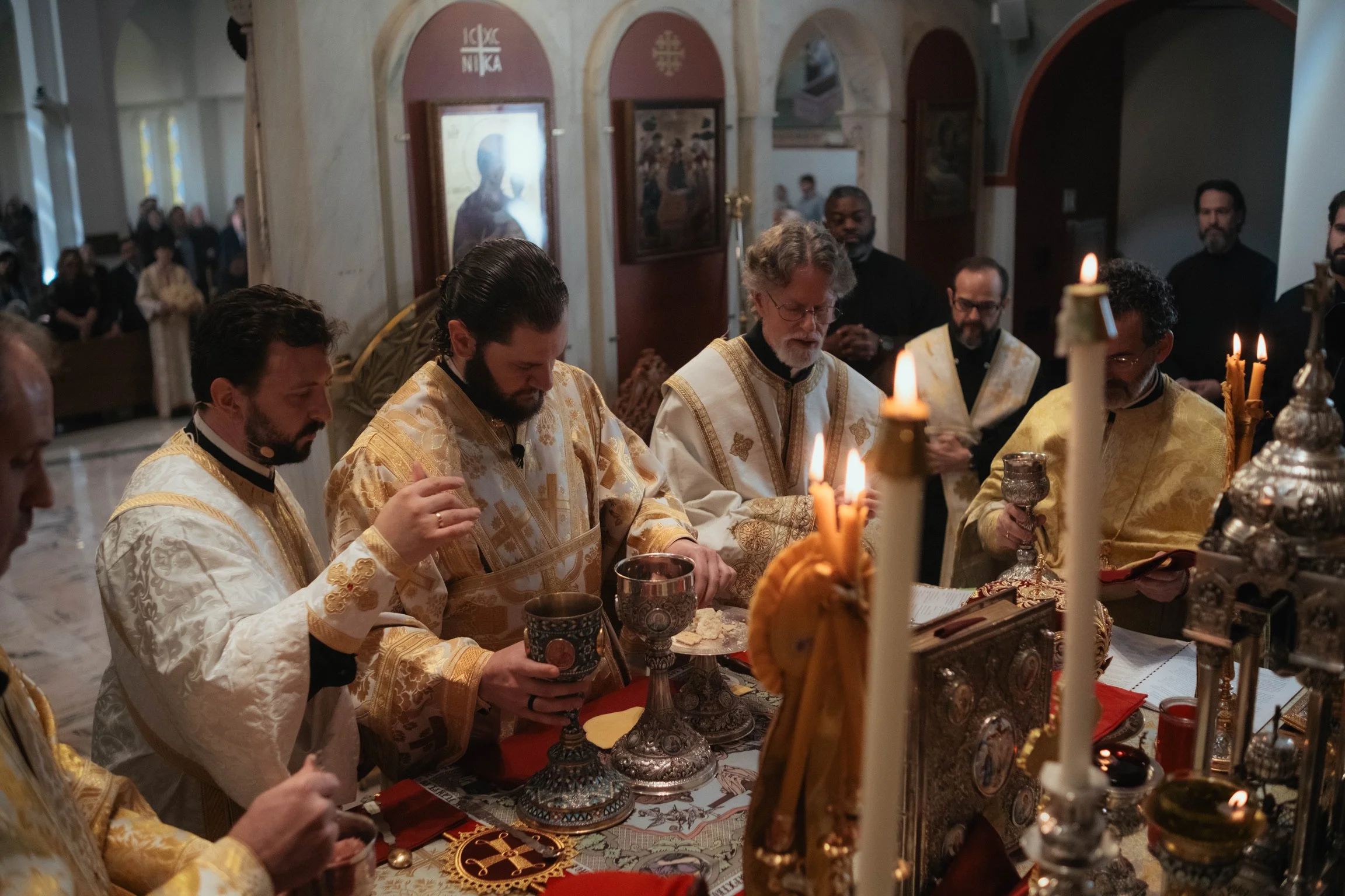Orthodox Christian priests and clergy participating in religious service, wearing gold vestments, holding chalices, with lit candles on an ornate altar in a church interior.