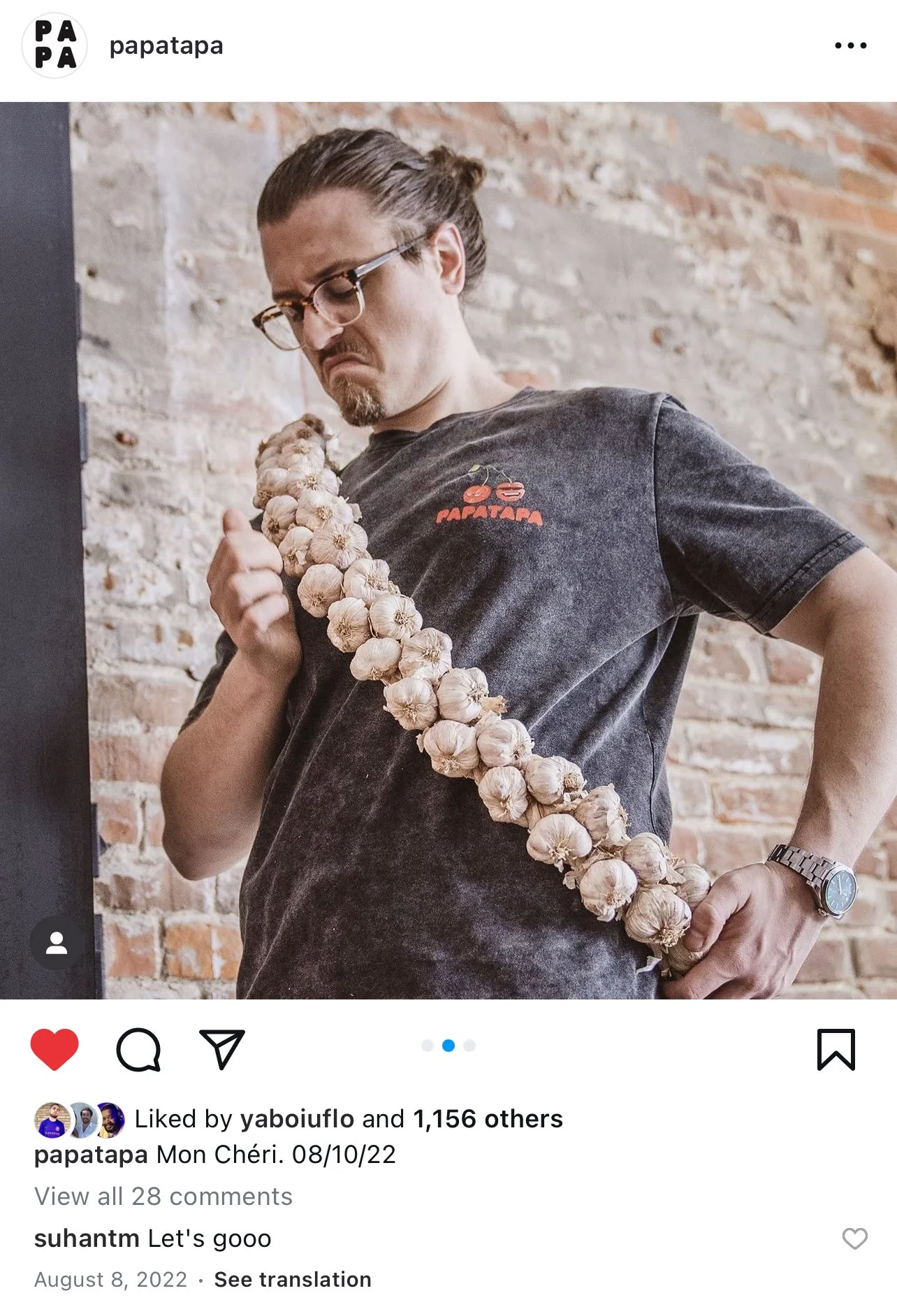 A man with glasses and a man bun hairstyle holding a large bunch of garlic over his shoulder, standing in front of a brick wall.