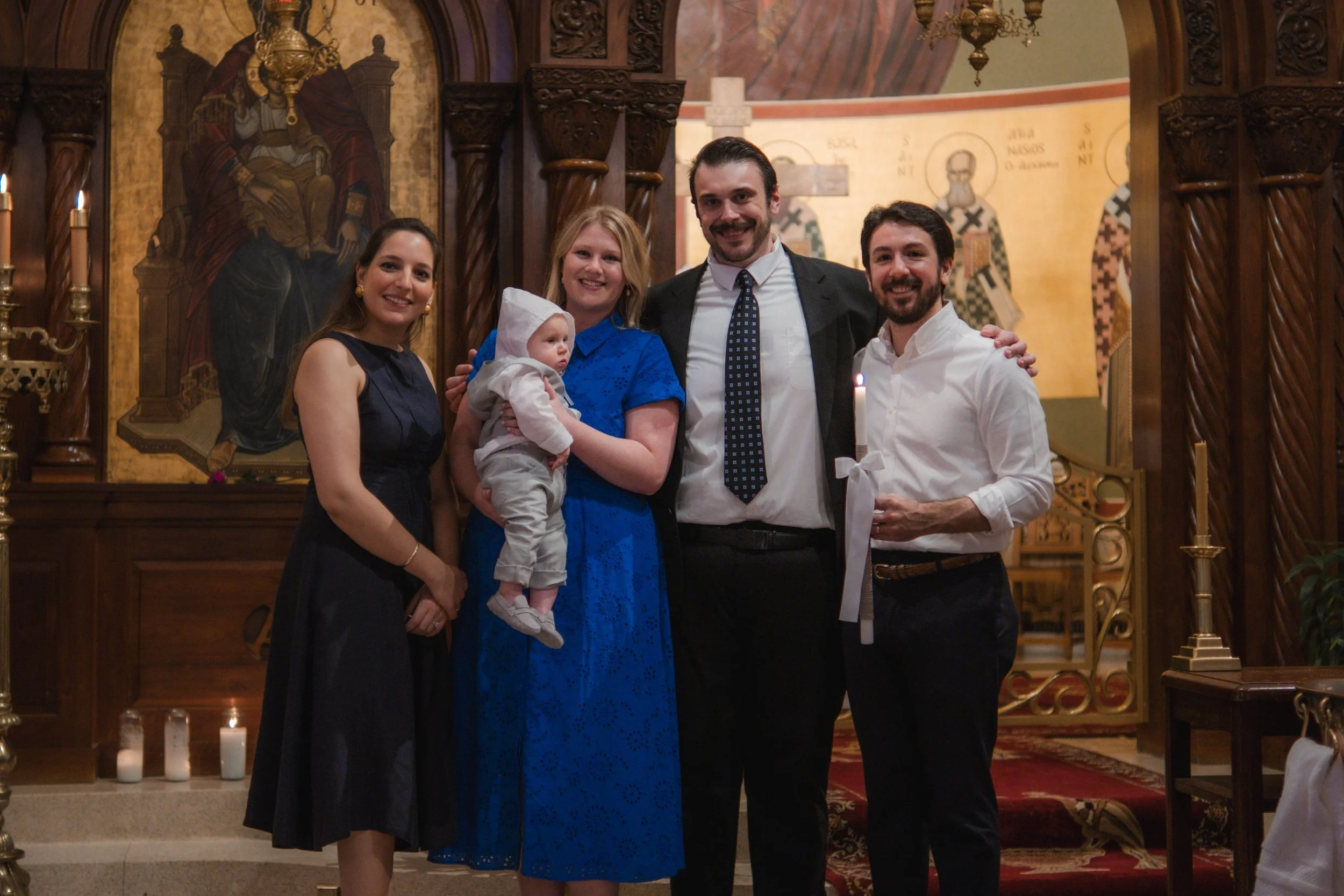 Group of five people standing inside a church, celebrating a baptism or christening, with a priest or officiant holding a lit candle. One woman holds a baby, with ornate religious icons and candles in the background.