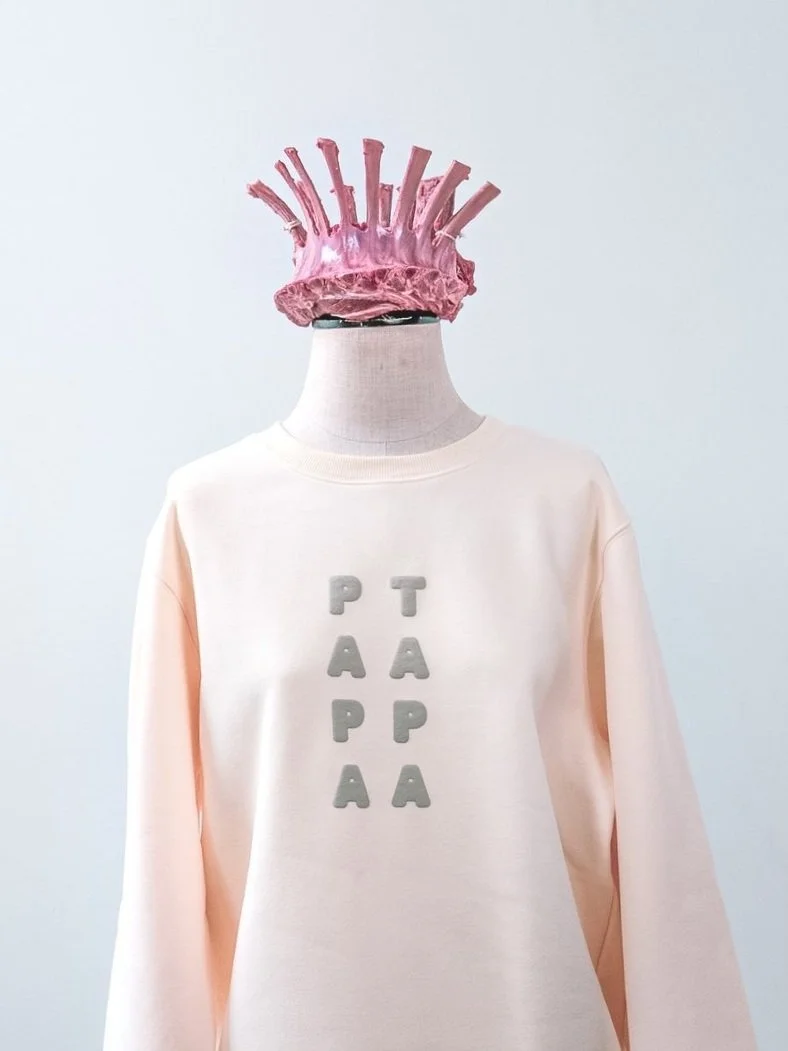 A mannequin wearing a pink headpiece resembling the Statue of Liberty's crown and a light pink sweatshirt with the word "PEACE" printed vertically in gray letters.