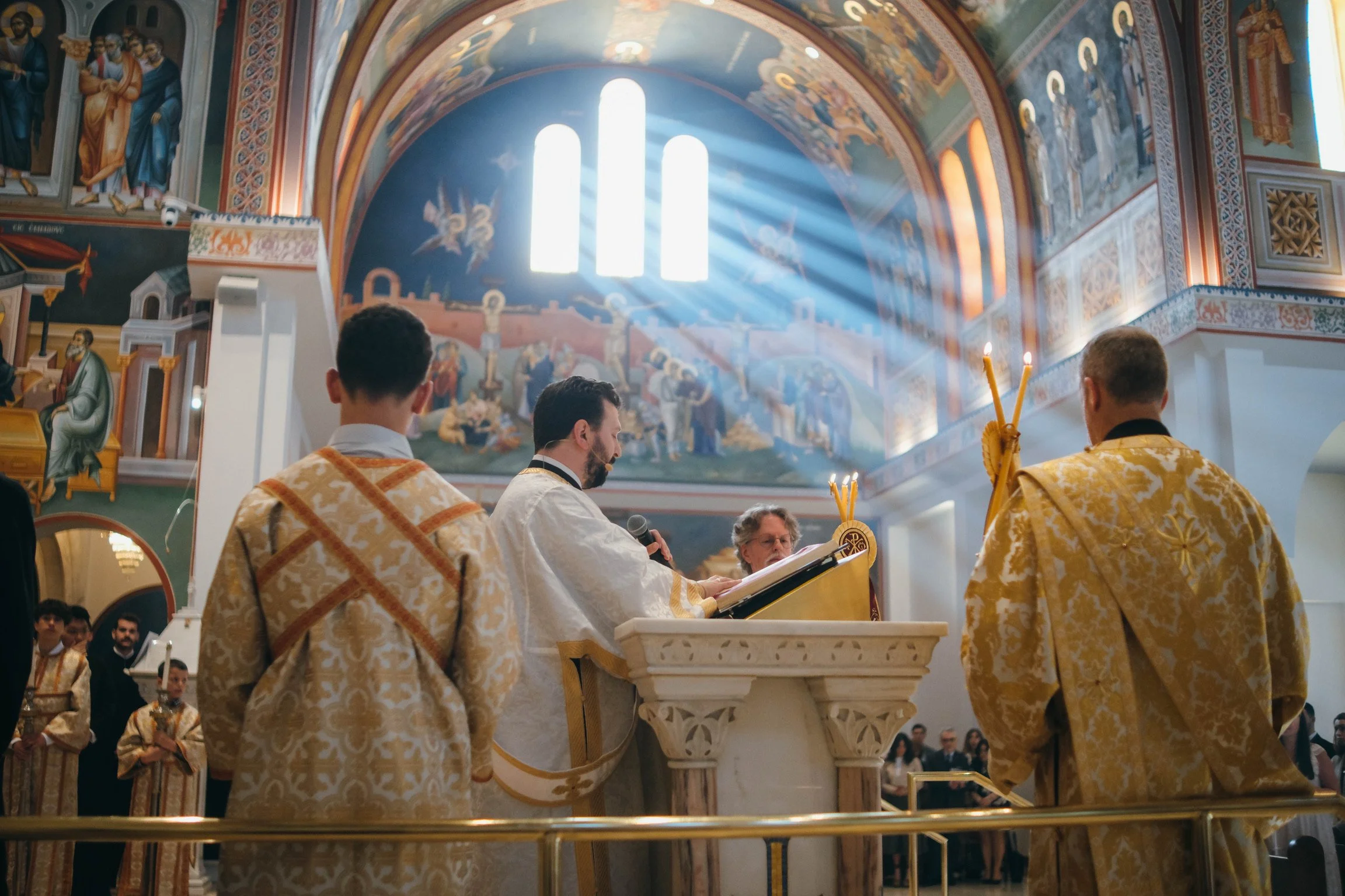 People participating in a religious ceremony inside an ornate church with colorful murals and sunlight streaming through stained glass windows.