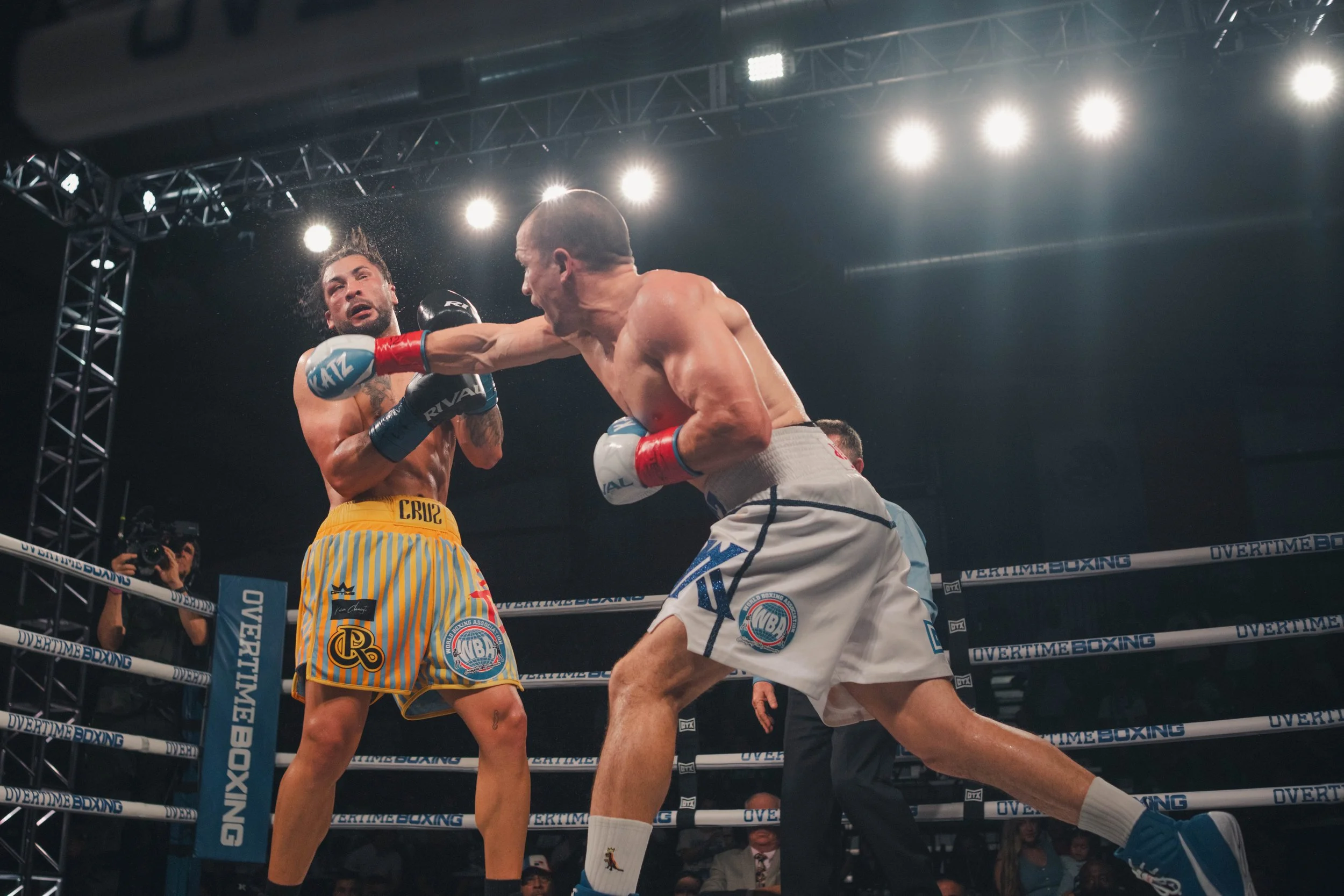 Two male boxers in a ring, one delivering a punch to the other's face. The boxer on the left is wearing yellow striped shorts, while the boxer on the right is in gray shorts. Bright overhead lights illuminate the scene, with a cameraman capturing the fight in the background.