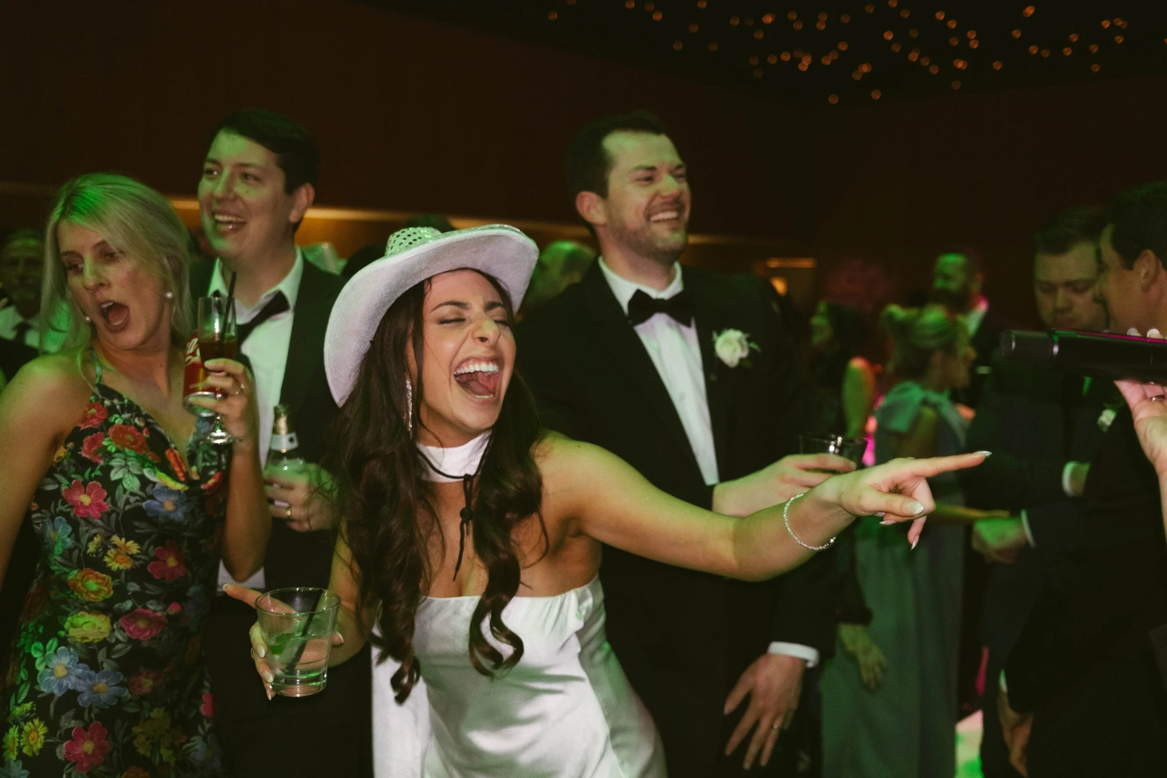 Group of people at a celebration, with women in colorful dresses and men in tuxedos, some holding drinks and smiling or laughing, with one woman pointing.