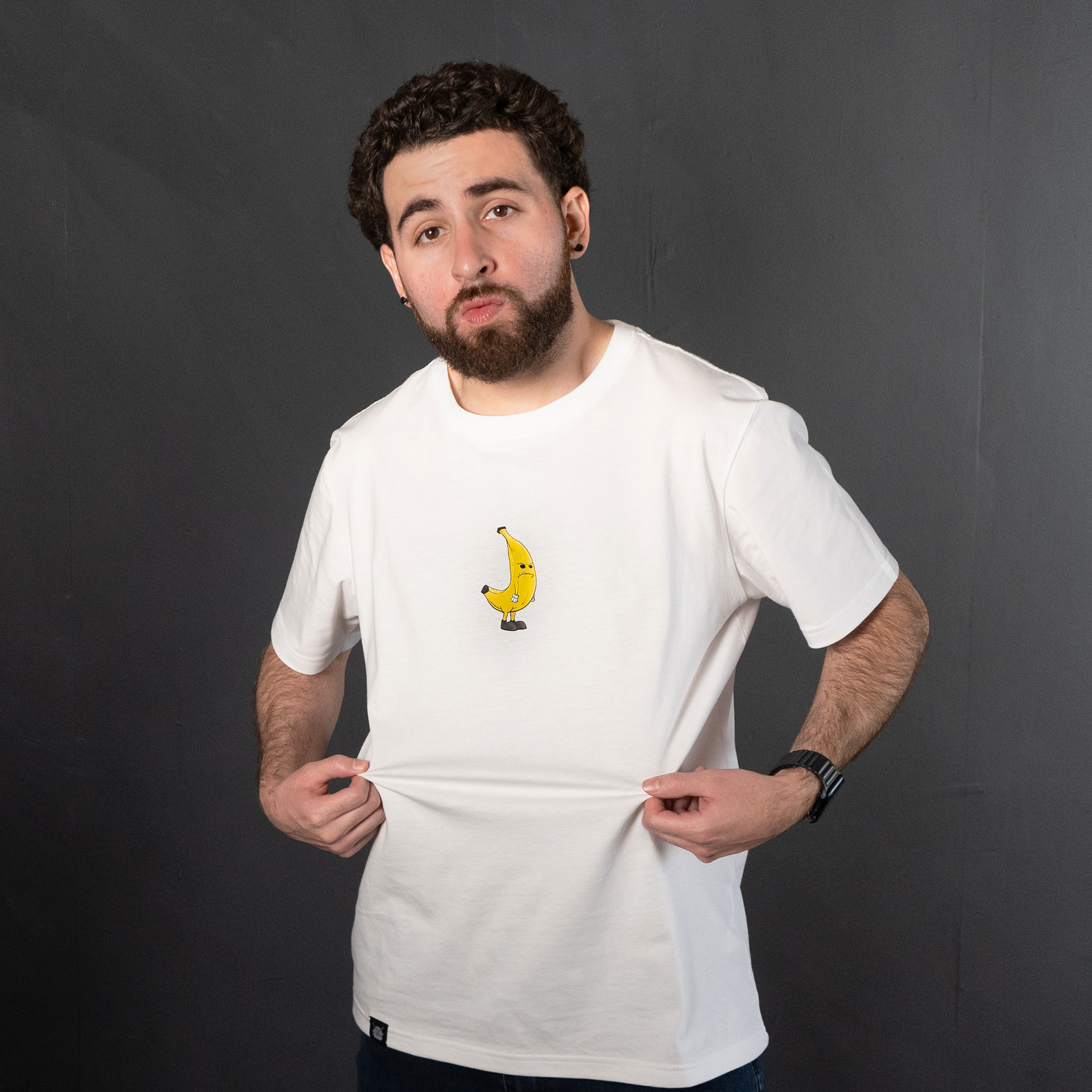 A man with curly hair and a beard, wearing a white T-shirt with a cartoon banana on it, pulls the shirt down slightly while standing against a dark gray background.
