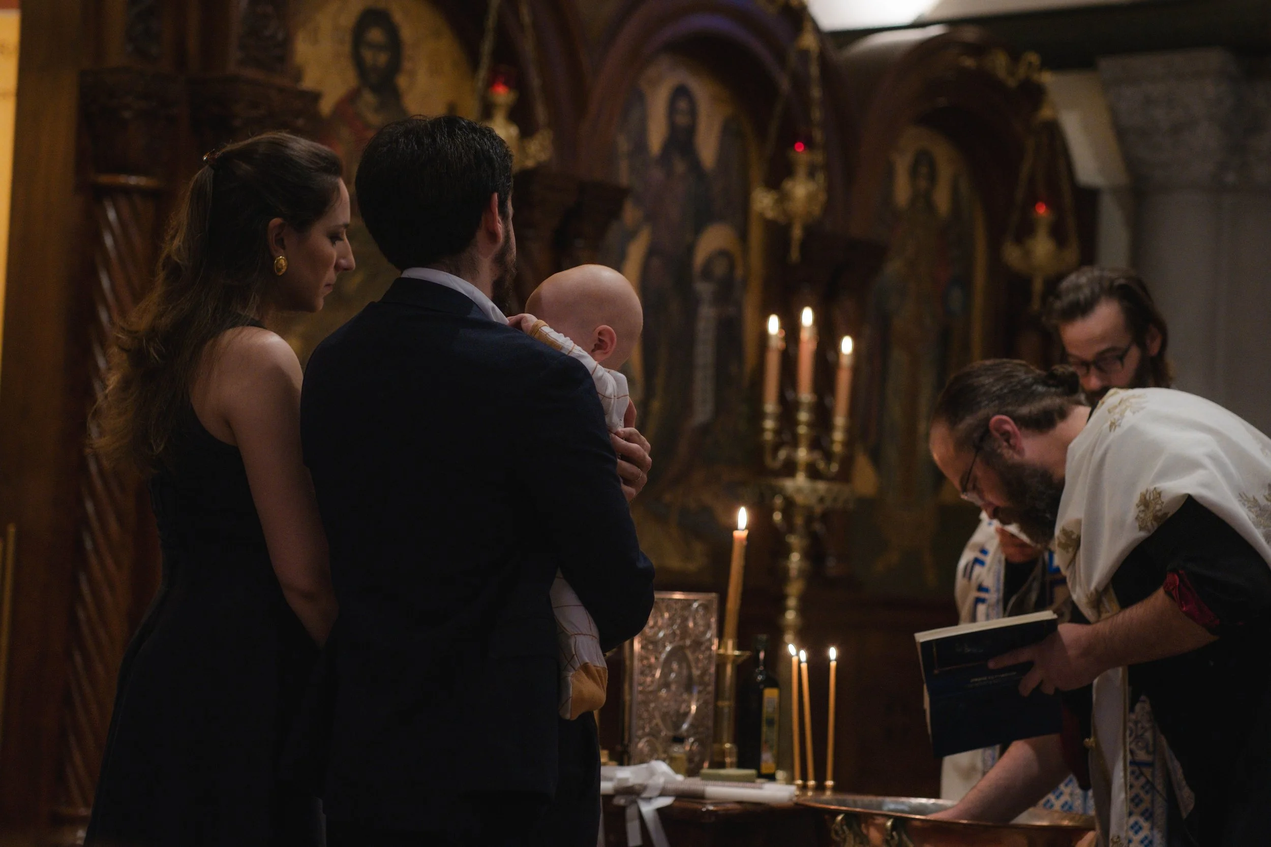 A religious baptism ceremony in a church with five people standing around an altar with lit candles, a priest performing the ritual, and an icon of Jesus Christ in the background.