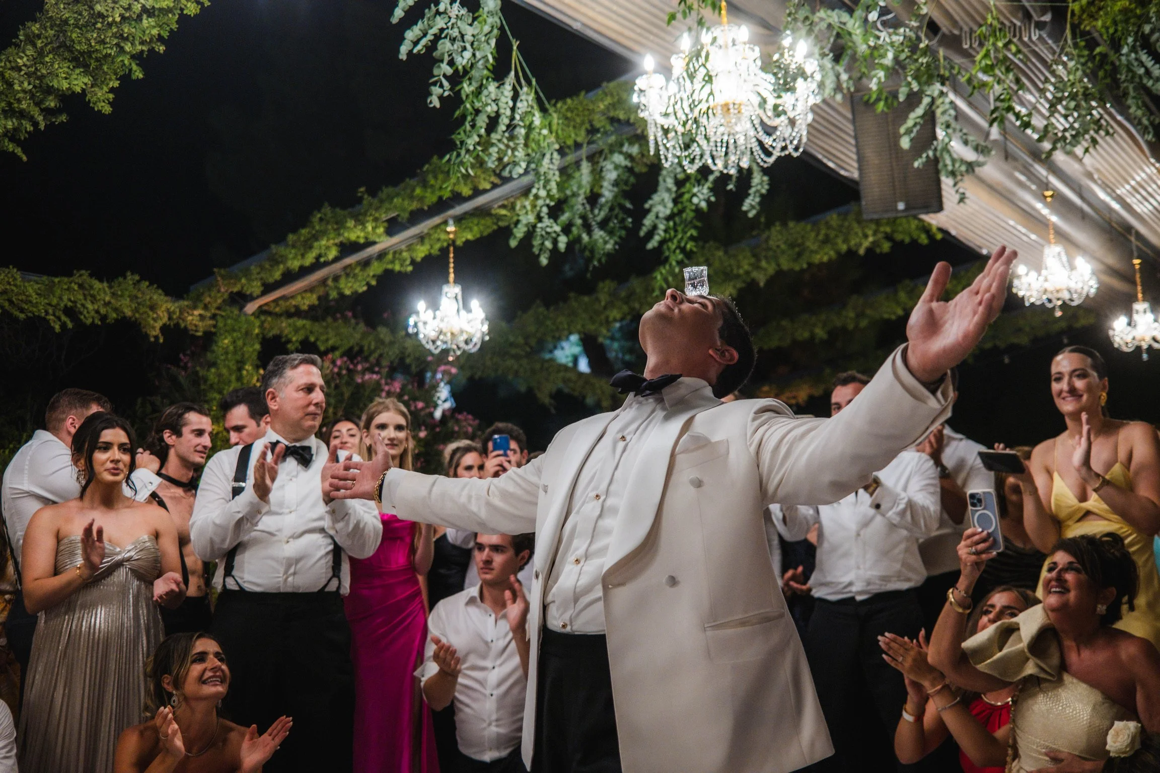 A man in a white tuxedo with a black bow tie balances a glass of water on his nose while standing with arms outstretched at a wedding reception. Surrounding guests are clapping and smiling under chandeliers and hanging greenery at night.