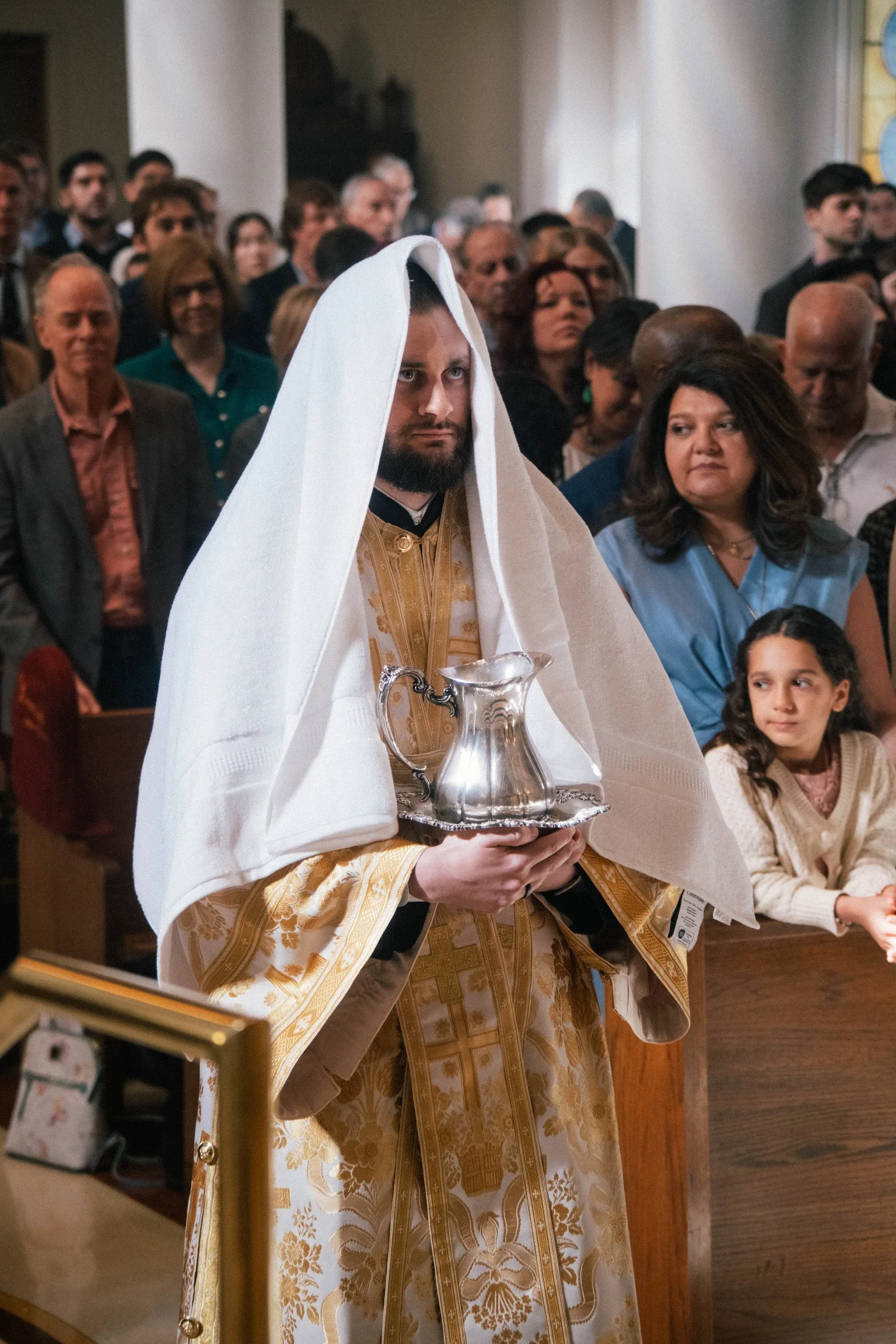 A priest holding a silver pitcher during a religious ceremony inside a church with a congregation of men, women, and children in the background.