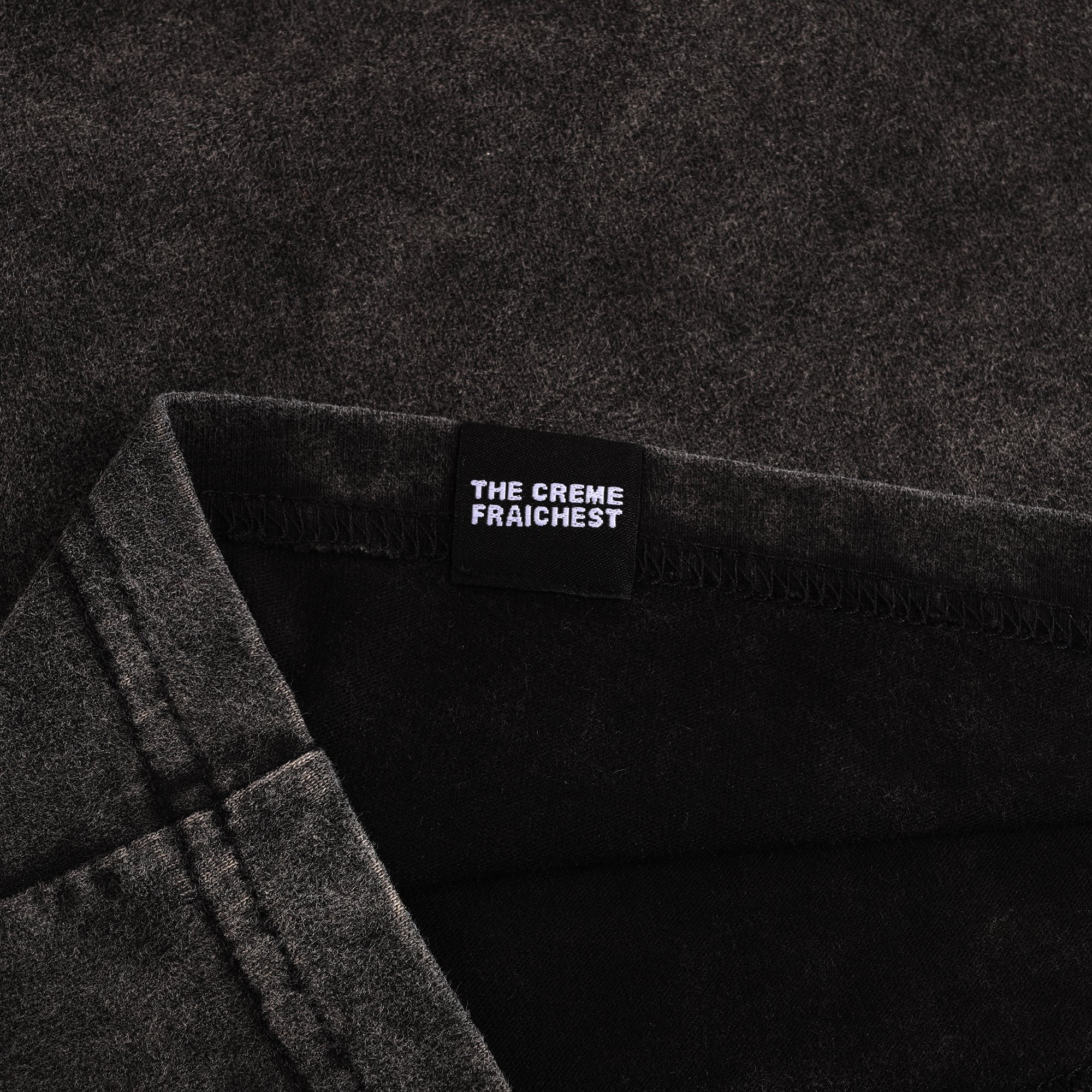 Close-up of a black clothing label that reads 'THE CREME FRAICHIEST' sewn into a dark gray fabric garment.