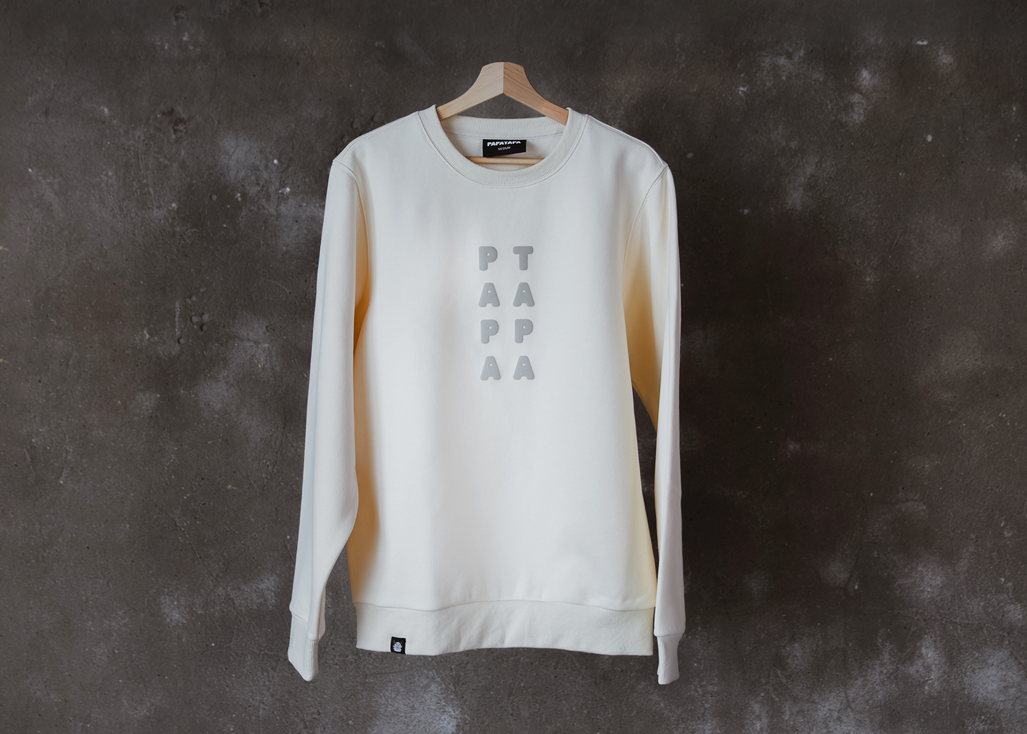White sweatshirt with the word 'PEACE' printed vertically in gray letters on the front, hanging on a wooden hanger against a dark textured wall.