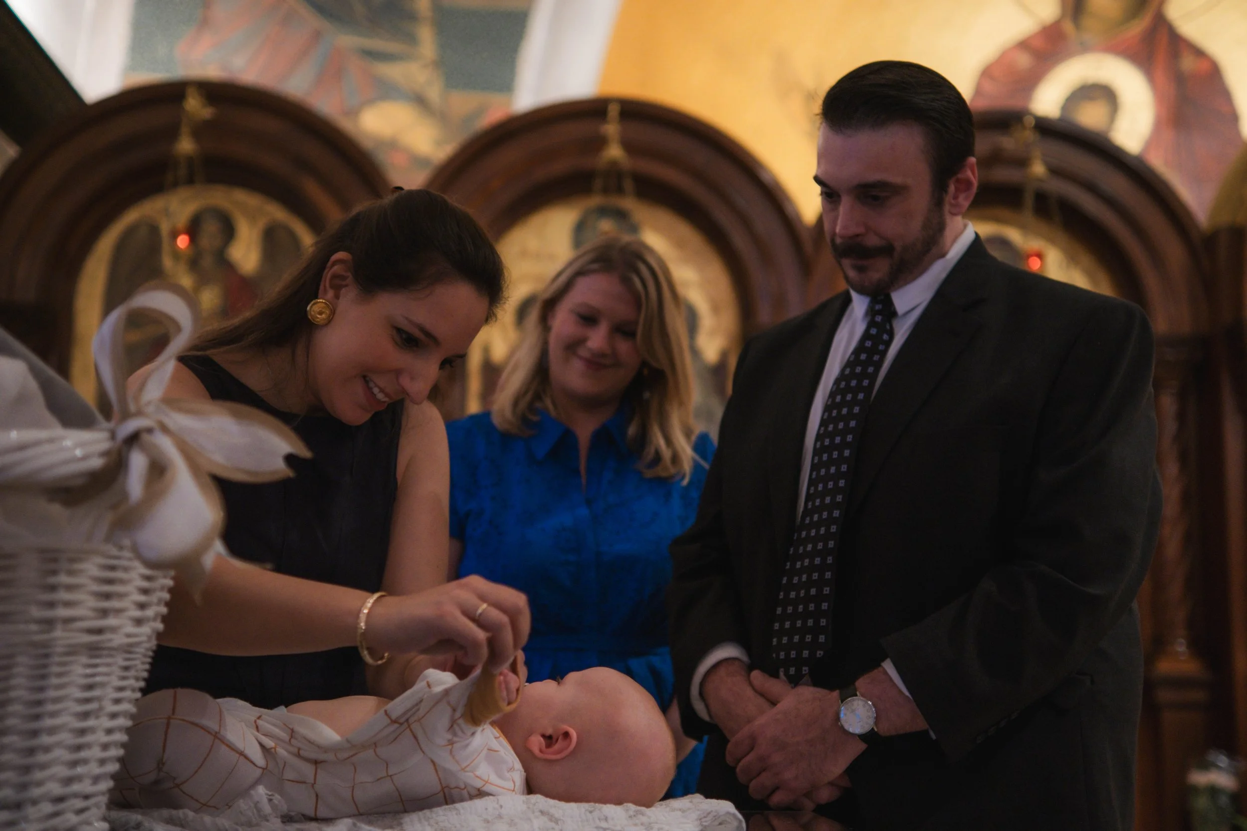 A baptism ceremony inside a church, with a woman holding a baby in a baptismal gown, surrounded by two women and a man, all smiling and dressed in formal attire.