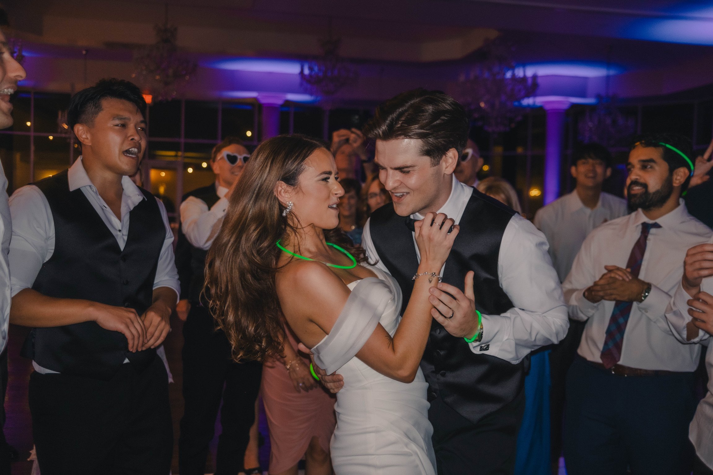 Couple dancing closely at a wedding reception surrounded by guests, with colorful lighting and chandeliers in the background.