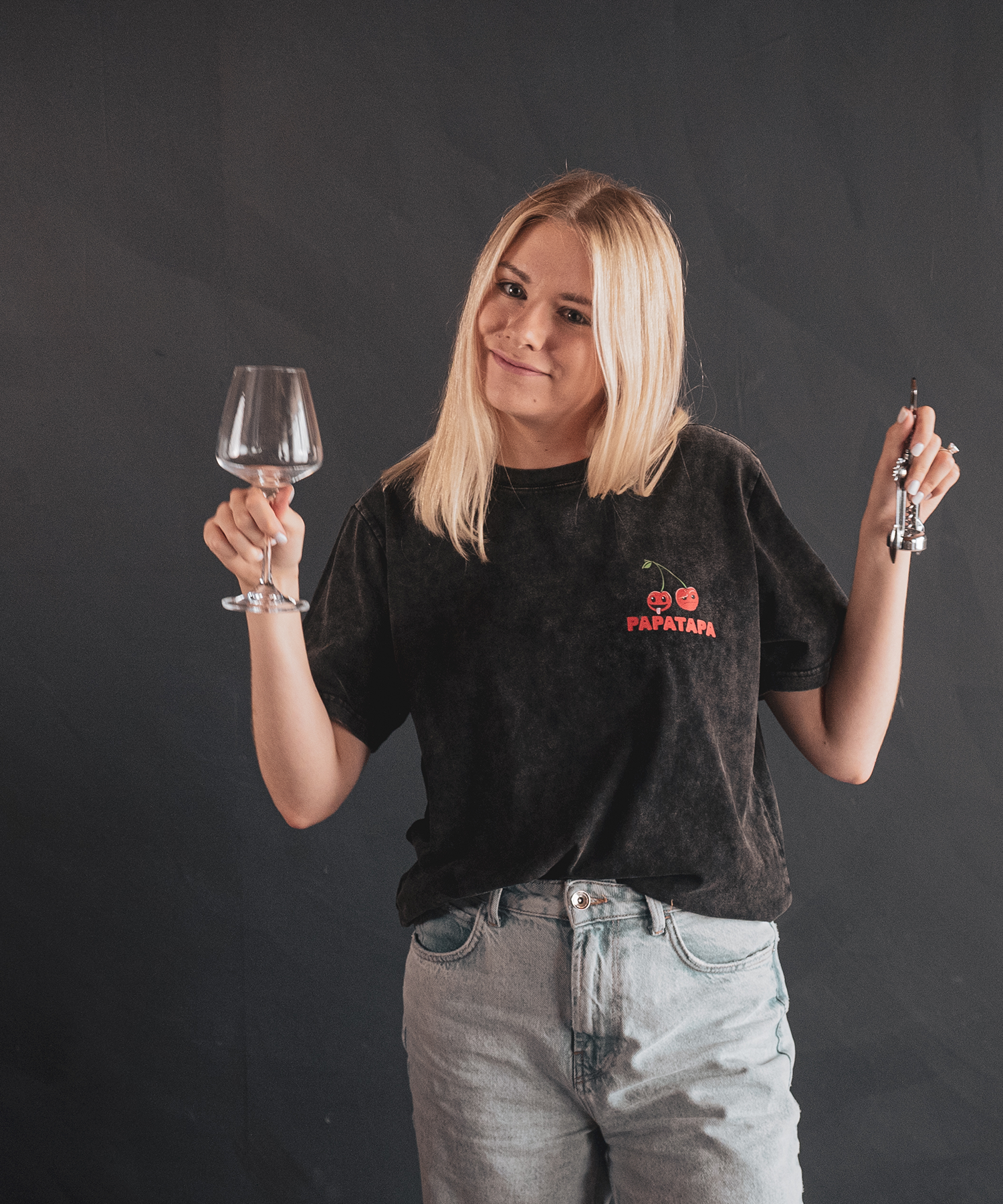 A young woman with blonde hair, wearing a black t-shirt with cherry graphics and the word 'PAPATAPA' on it, holding an empty wine glass in her right hand and a corkscrew in her left, standing against a dark background.