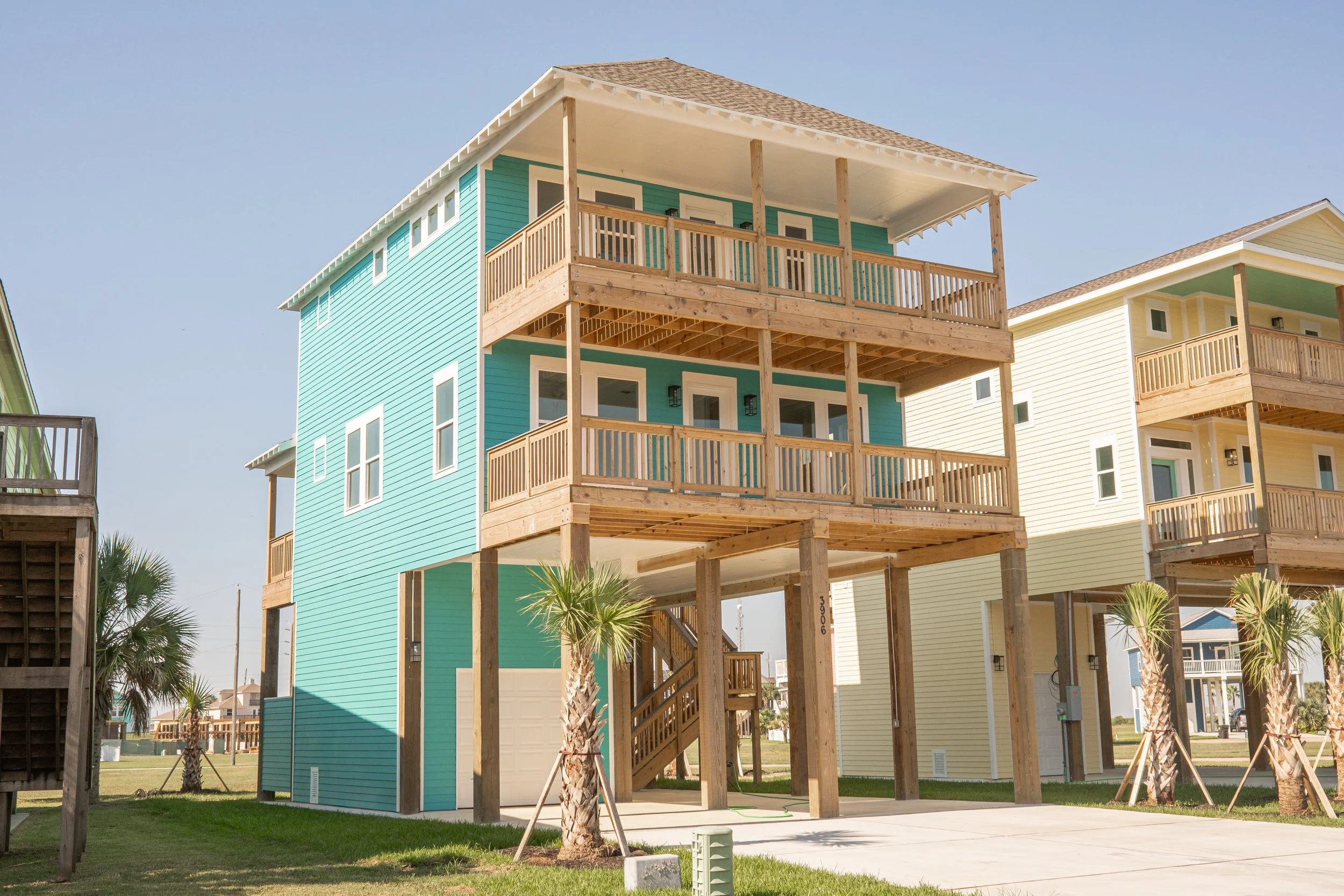 Colorful elevated beach house with teal siding, yellow siding, and wooden balconies supported by wooden posts, surrounded by small palm trees, in a sunny coastal neighborhood.