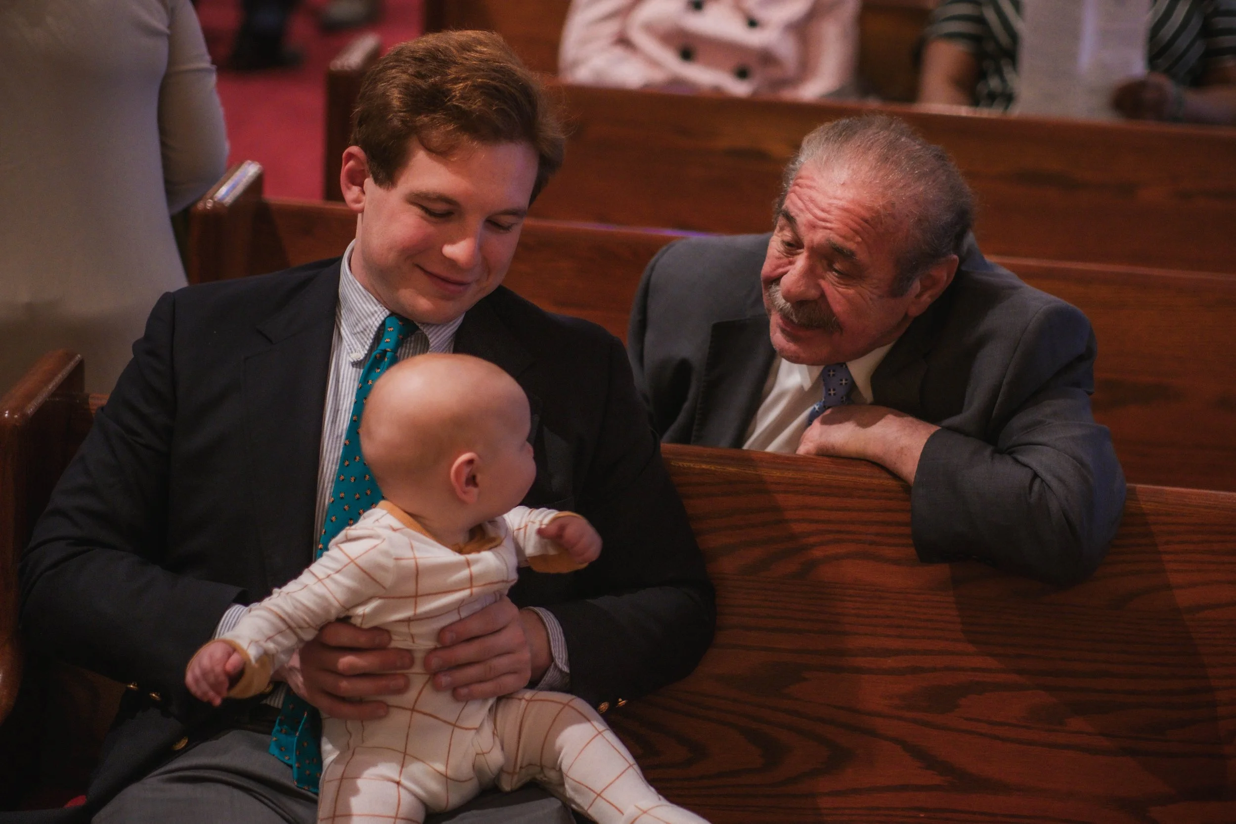 A young man in a suit holding a baby while smiling and looking at an older man, in a church with wooden pews.