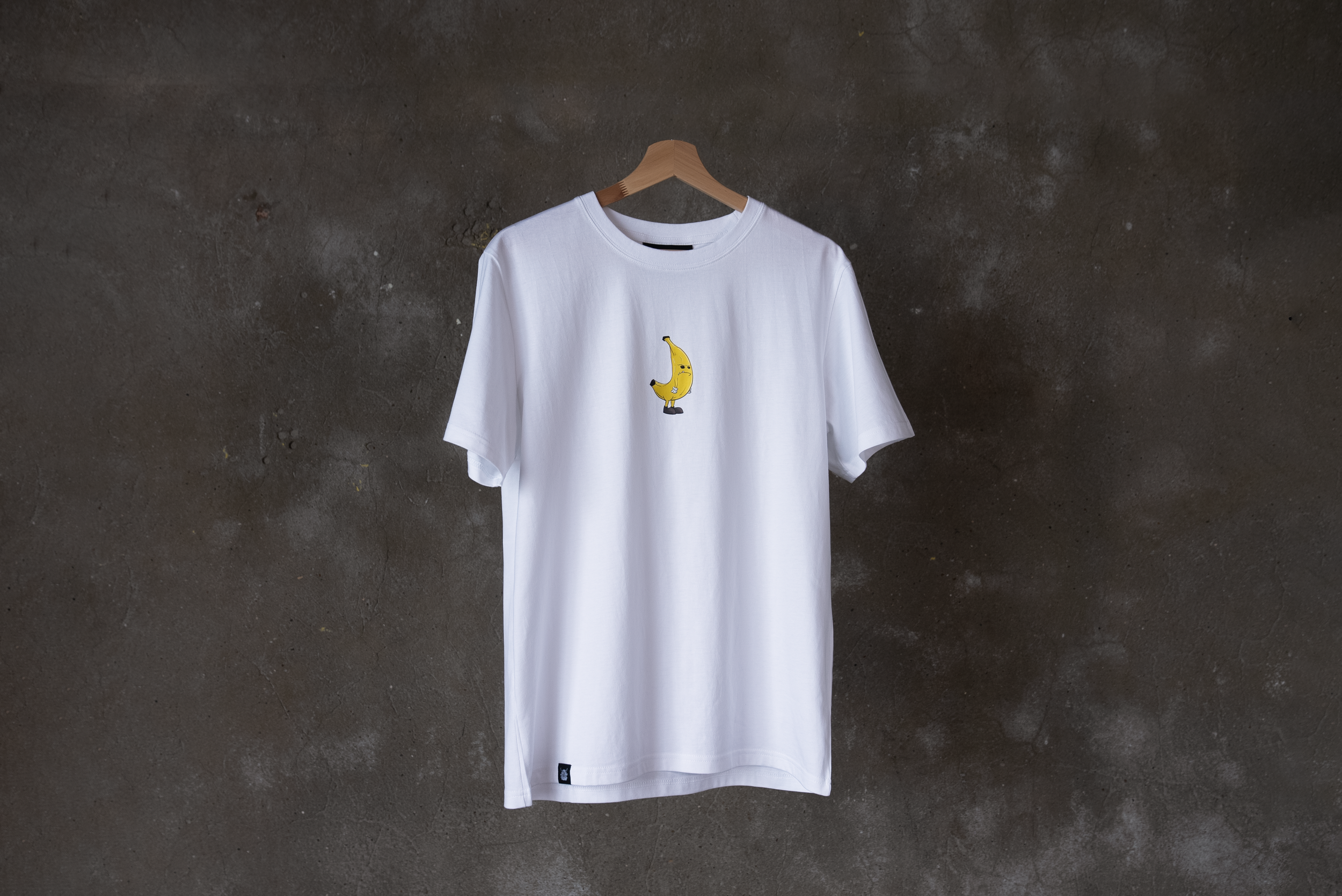 White T-shirt with a cartoon banana character printed on the front, hanging on a wooden hanger against a dark, textured concrete wall.