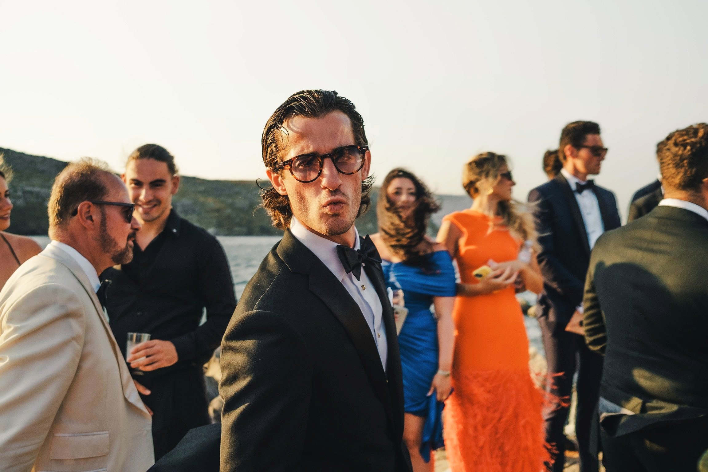 Group of people dressed in formal attire socializing outdoors during sunset near a body of water.