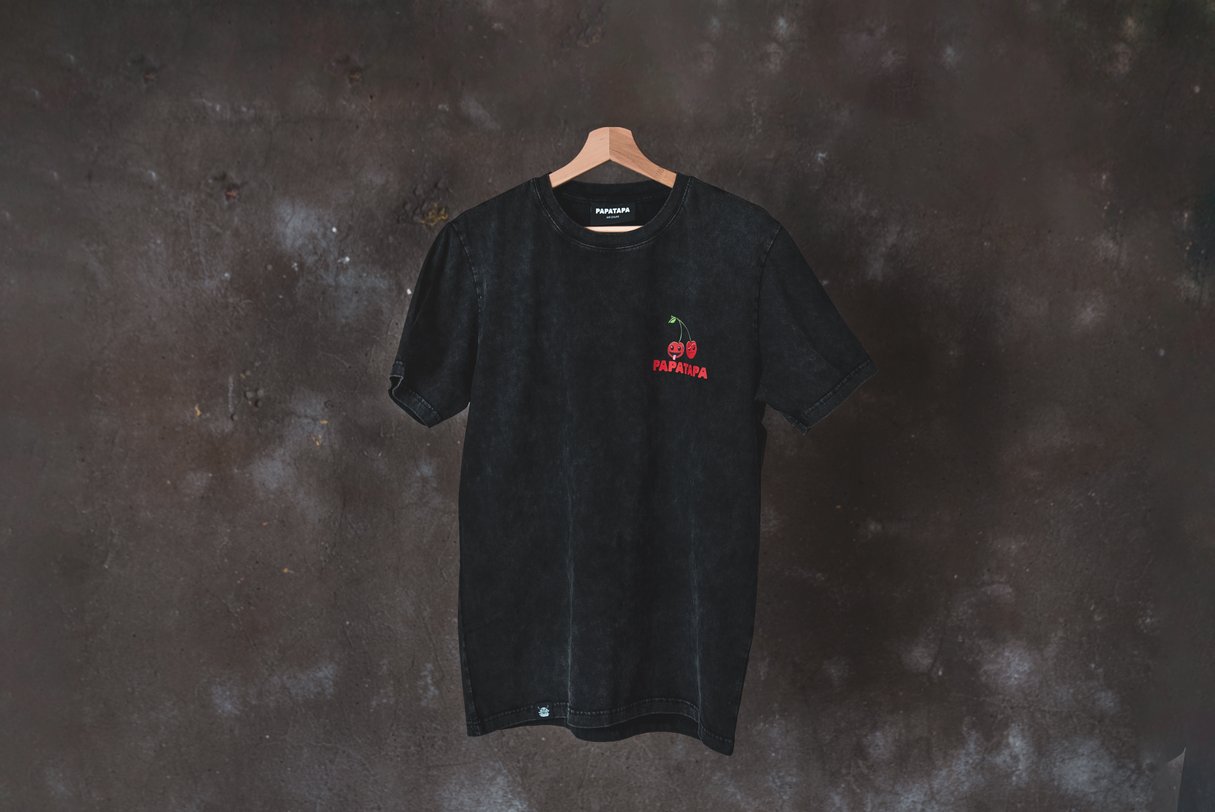 Black T-shirt with embroidered cherry graphic and red text saying 'PAPATAPA', hanging on a wooden hanger against a dark textured wall.