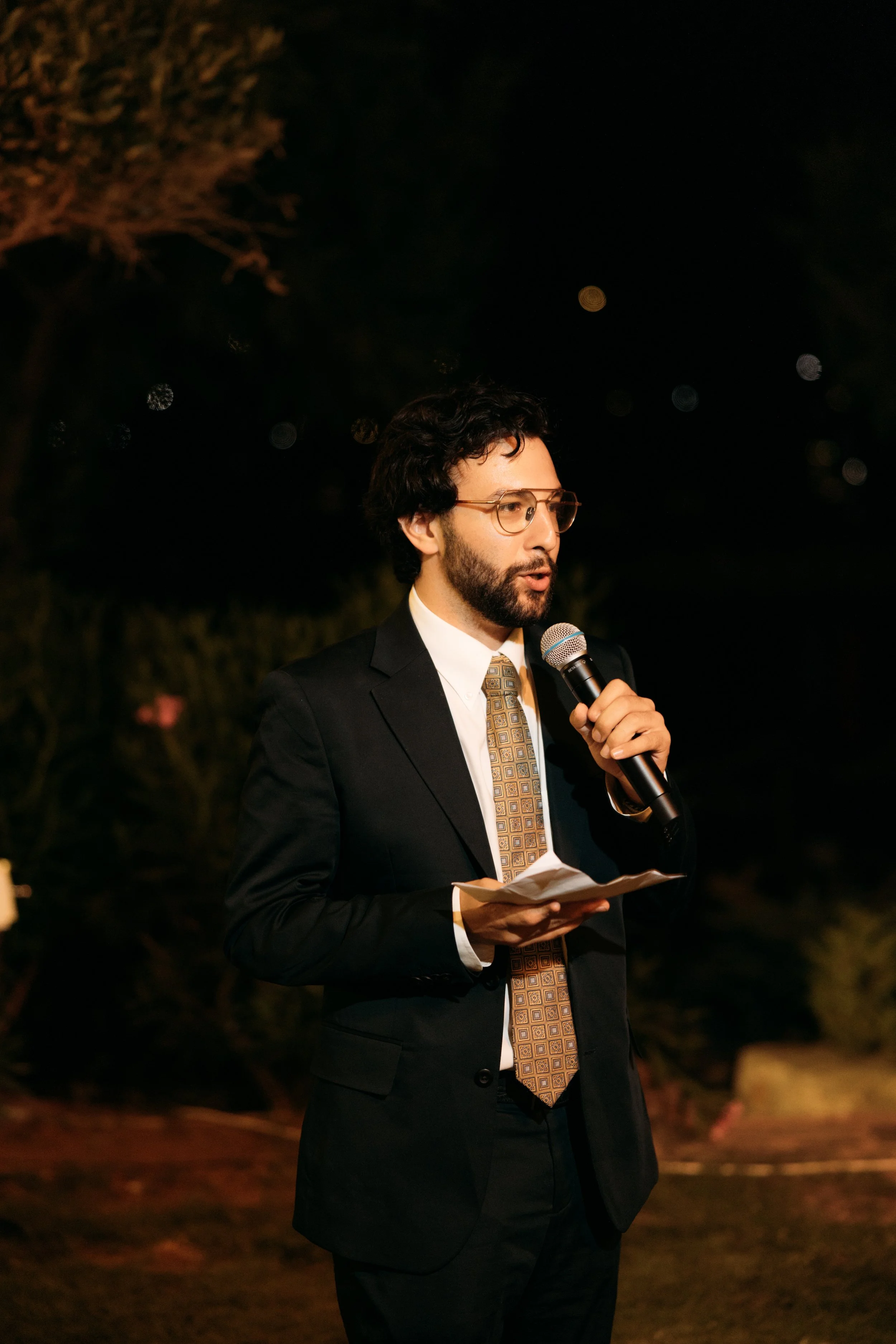Man in a suit holding a microphone and reading from papers, speaking at an outdoor nighttime event.