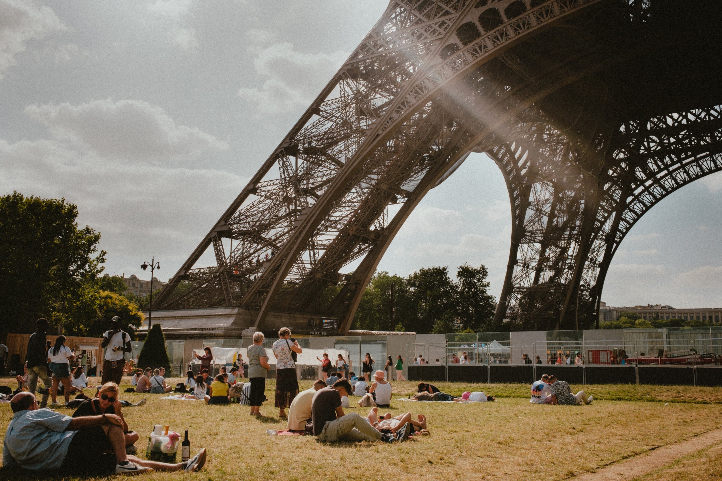 People sitting on the grass and walking near the Eiffel Tower with a construction fencing and trees in the background, under a partly cloudy sky.