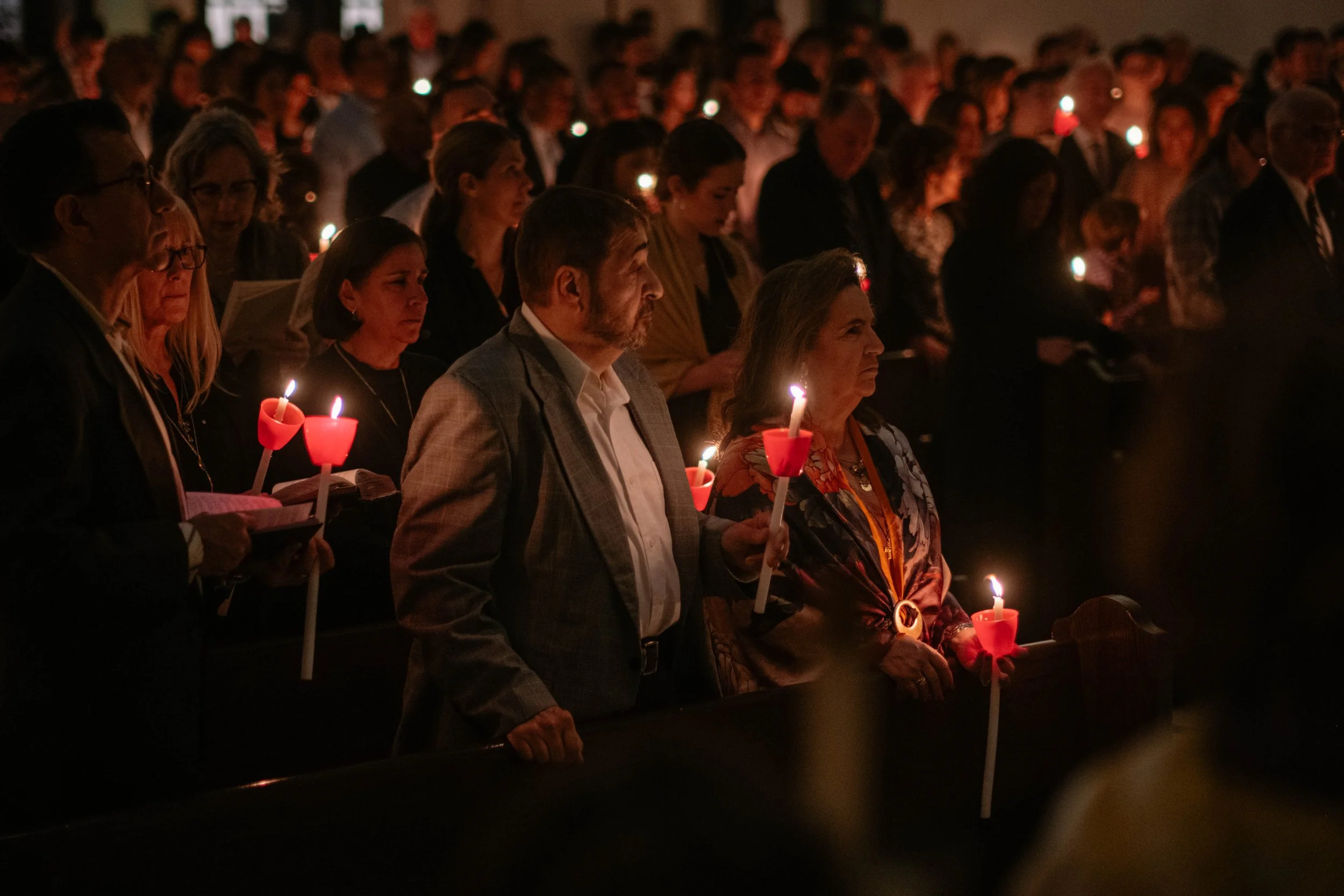 People participating in a candlelight vigil, holding candles with red holders, in a dark indoor setting.