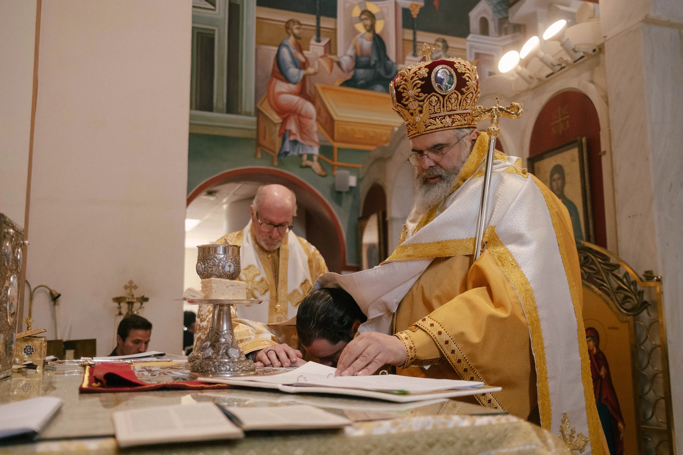 A religious ceremony taking place in an Orthodox church, featuring clergy in ornate vestments and a bishop wearing a gold crown. One clergy member is bowing with his head on the altar, while another looks down, with religious murals and icons visible