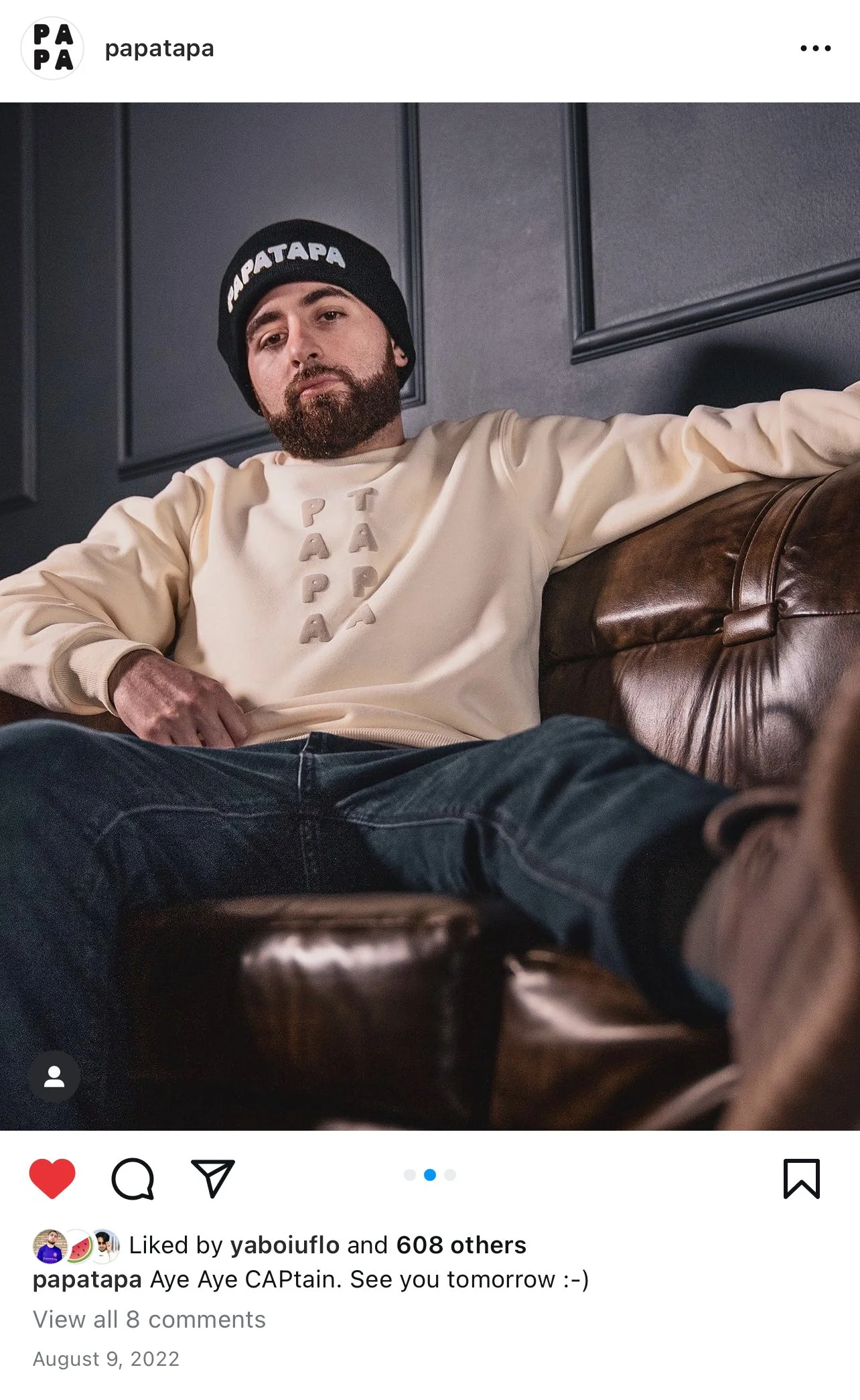 A young man with a beard wearing a black beanie and a cream-colored sweatshirt sitting on a brown leather couch against dark colored wall panels.