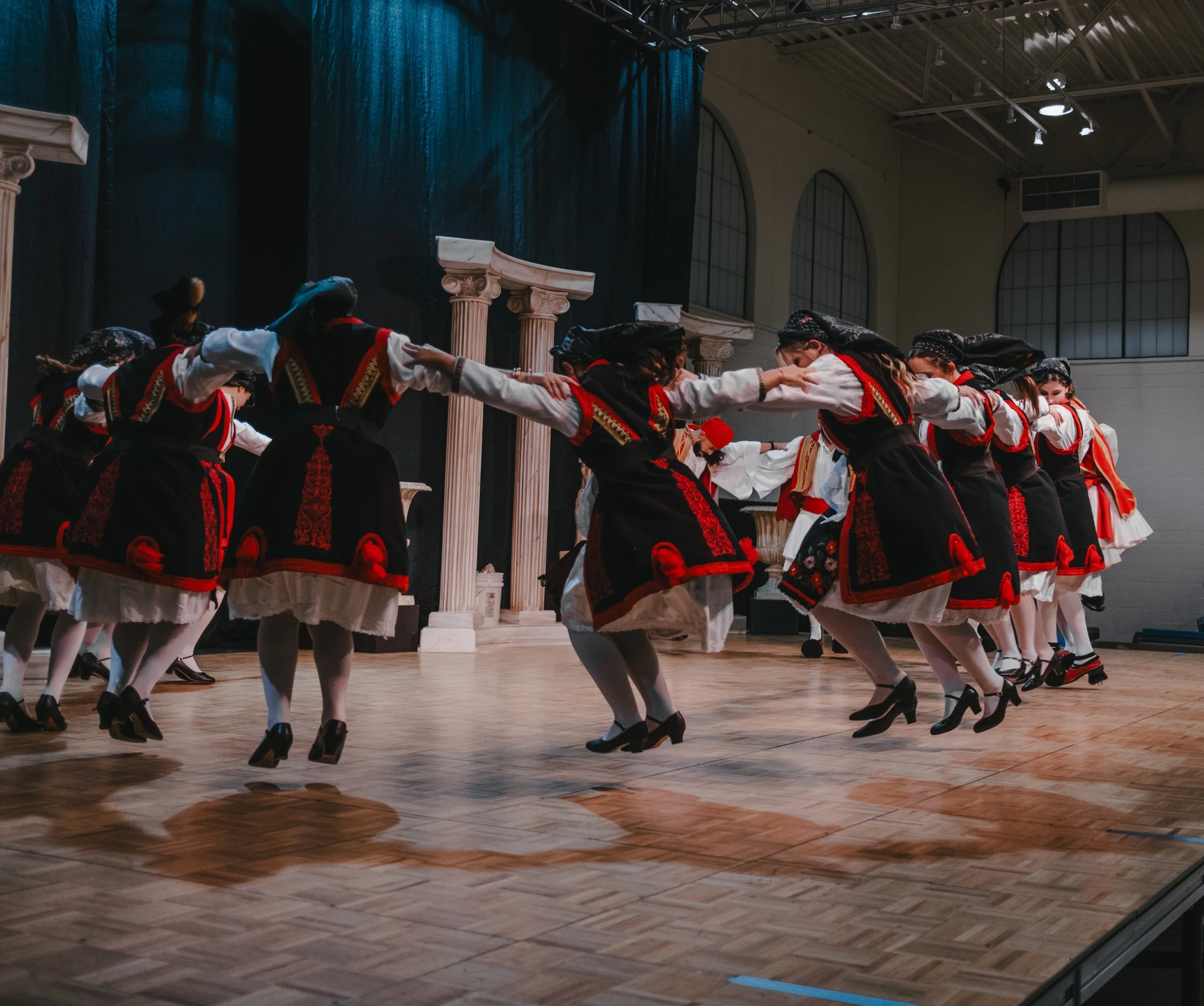 Women in traditional folk costumes performing a cultural dance on stage with Greek-style columns and arches in the background.
