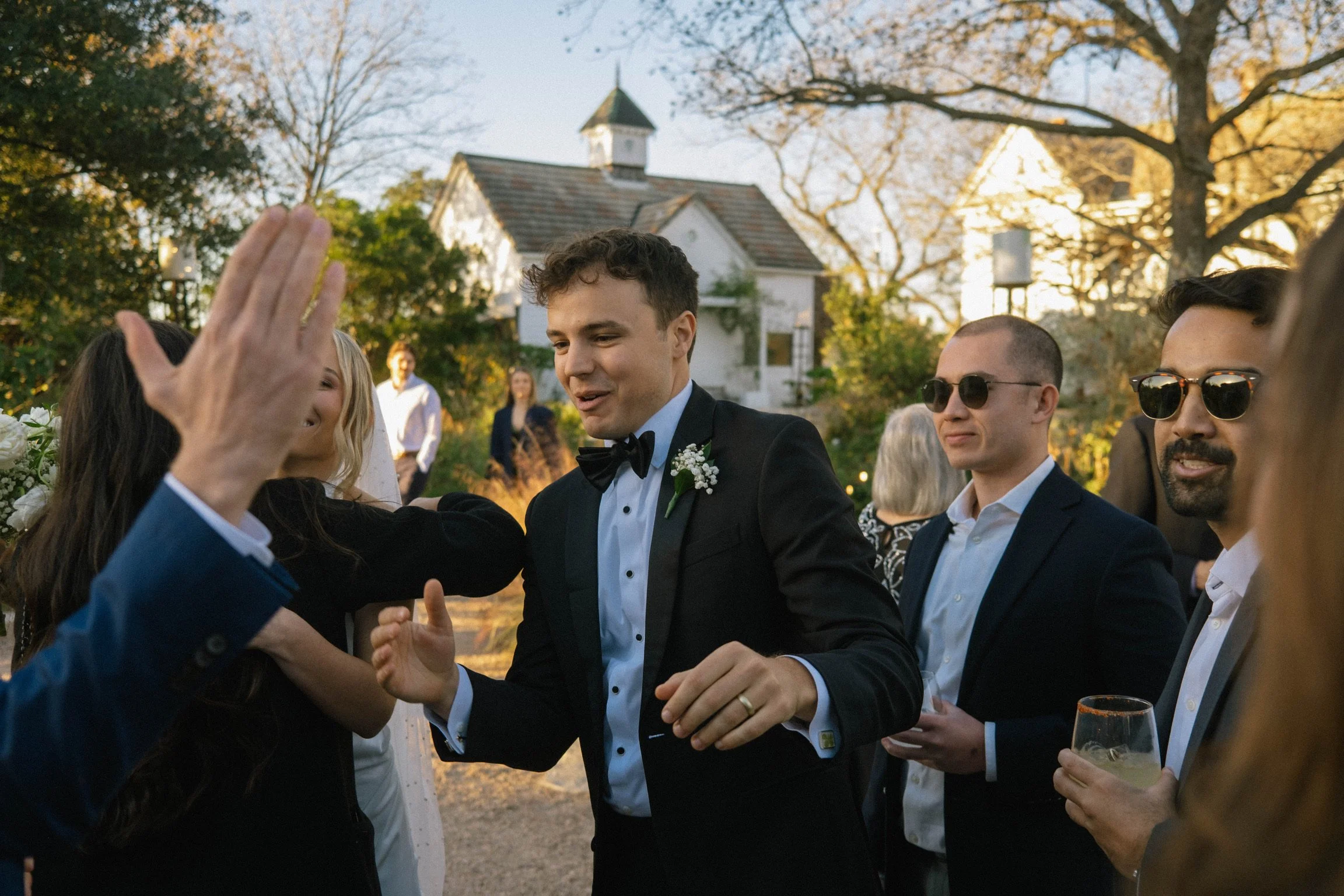 Smooth-skinned man in black tuxedo with bow tie, brushing off shoulder of woman in black dress, at outdoor wedding reception during sunset with guests and a white church in background.