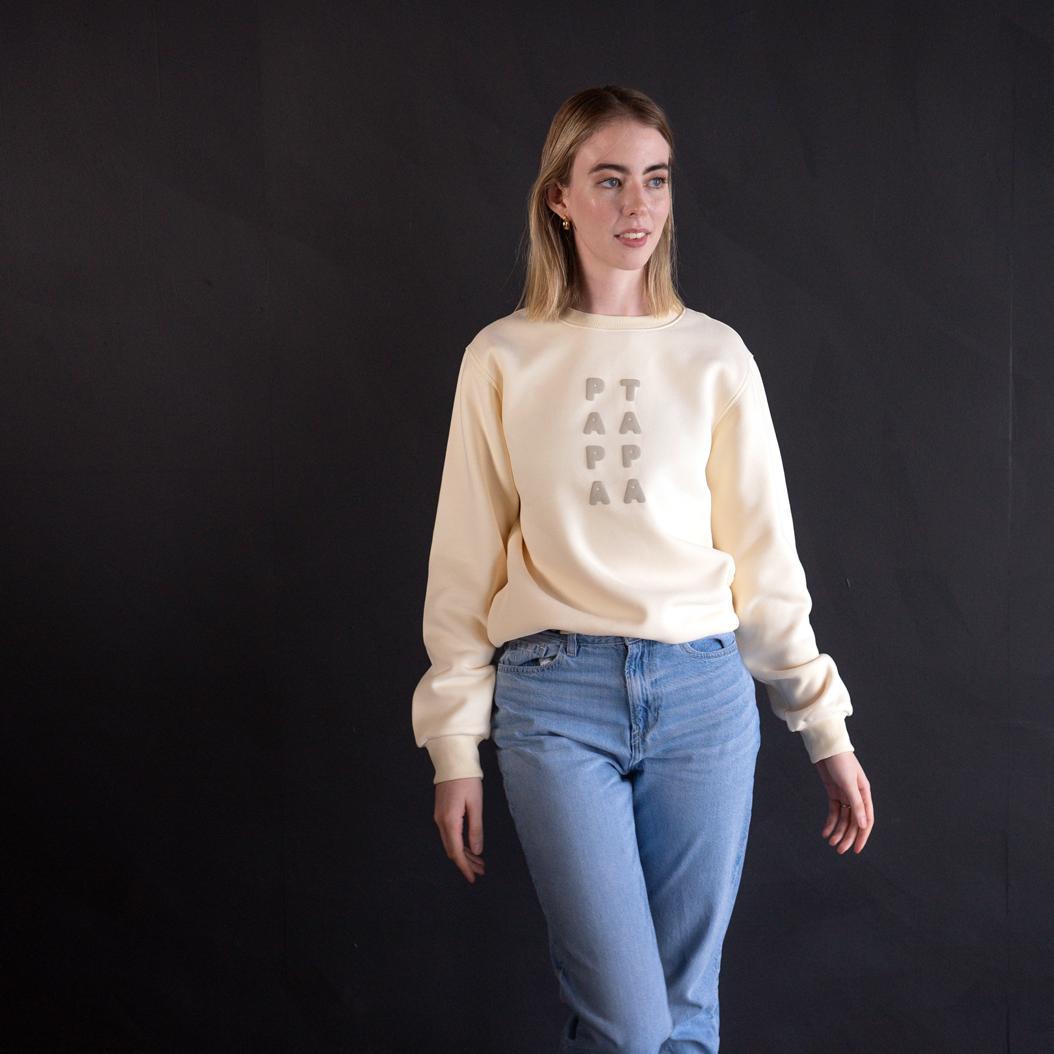 A young woman with blonde hair wearing a cream-colored sweatshirt with the words "PTA PAA" written vertically on the front and light blue jeans standing against a dark background.