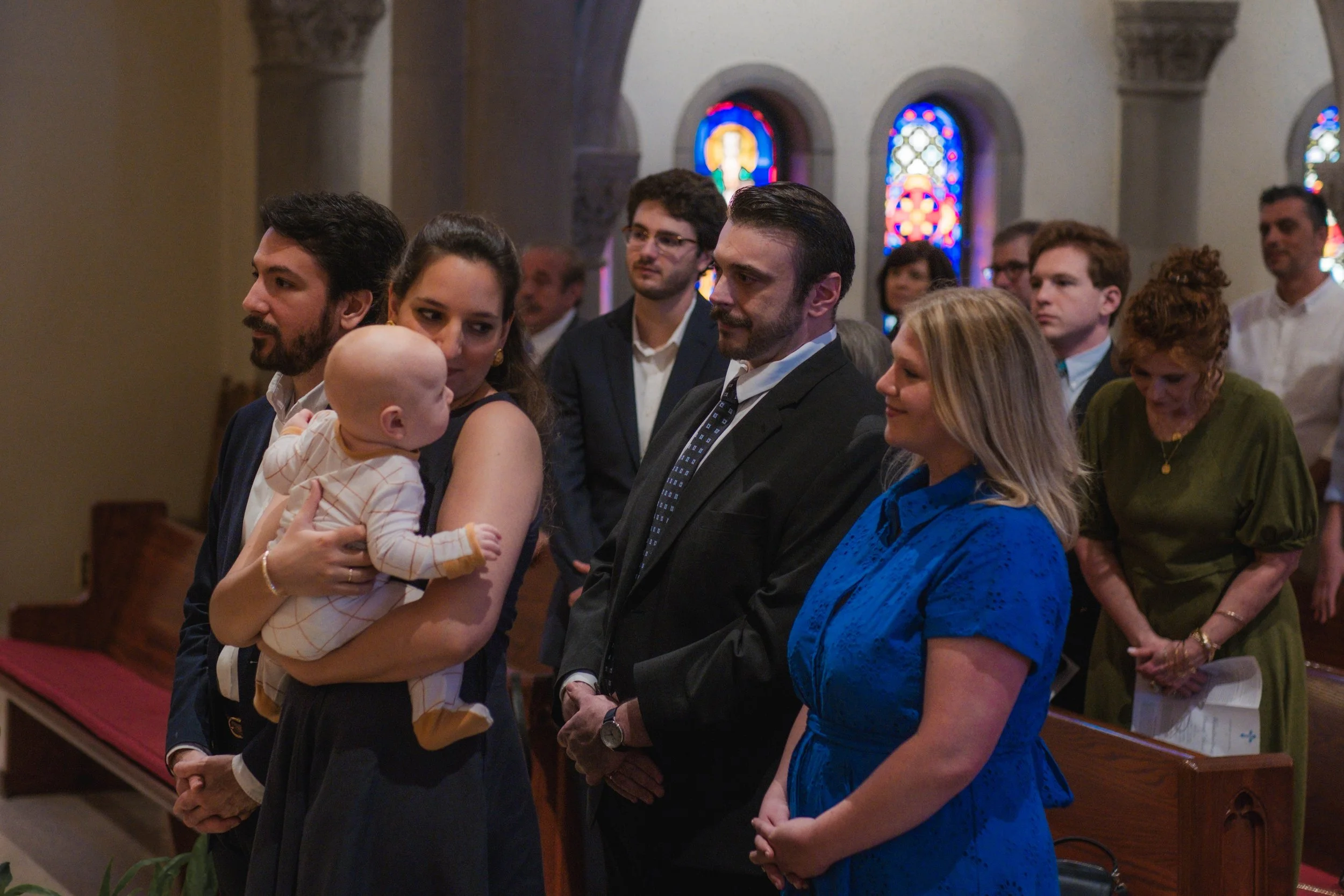 People attending a religious ceremony in a church, with stained glass windows in the background, including a woman holding a baby and others standing near pews.
