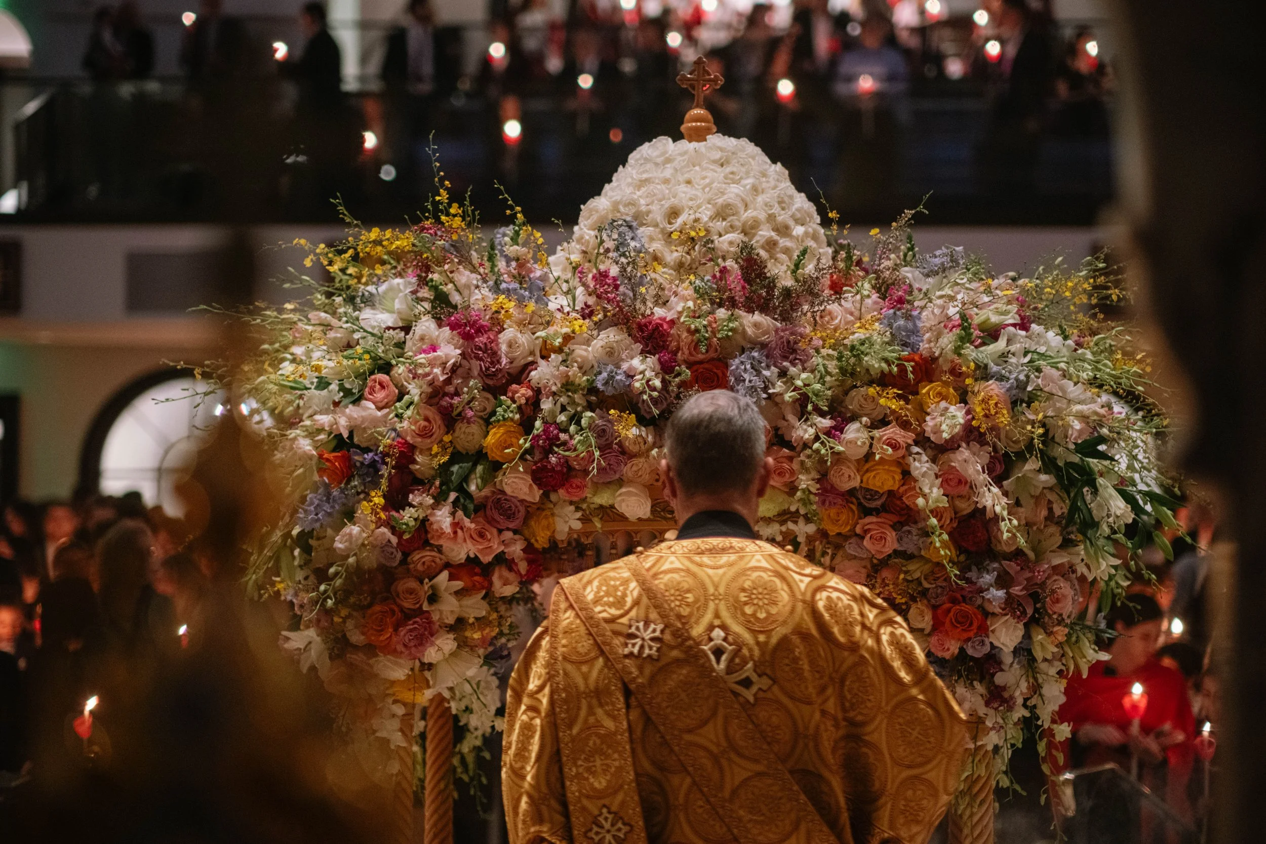 A religious clergy member, dressed in ornate gold vestments, faces an elaborate floral arrangement with white roses and various other colorful flowers during a ceremony in a church filled with people.