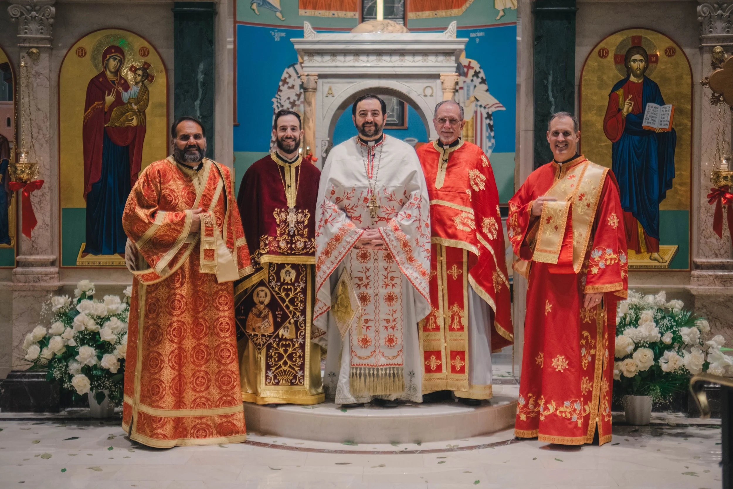 Six clergymen in ornate robes standing in front of an iconostasis in an Orthodox church, with icons of the Virgin Mary and Jesus Christ in the background.