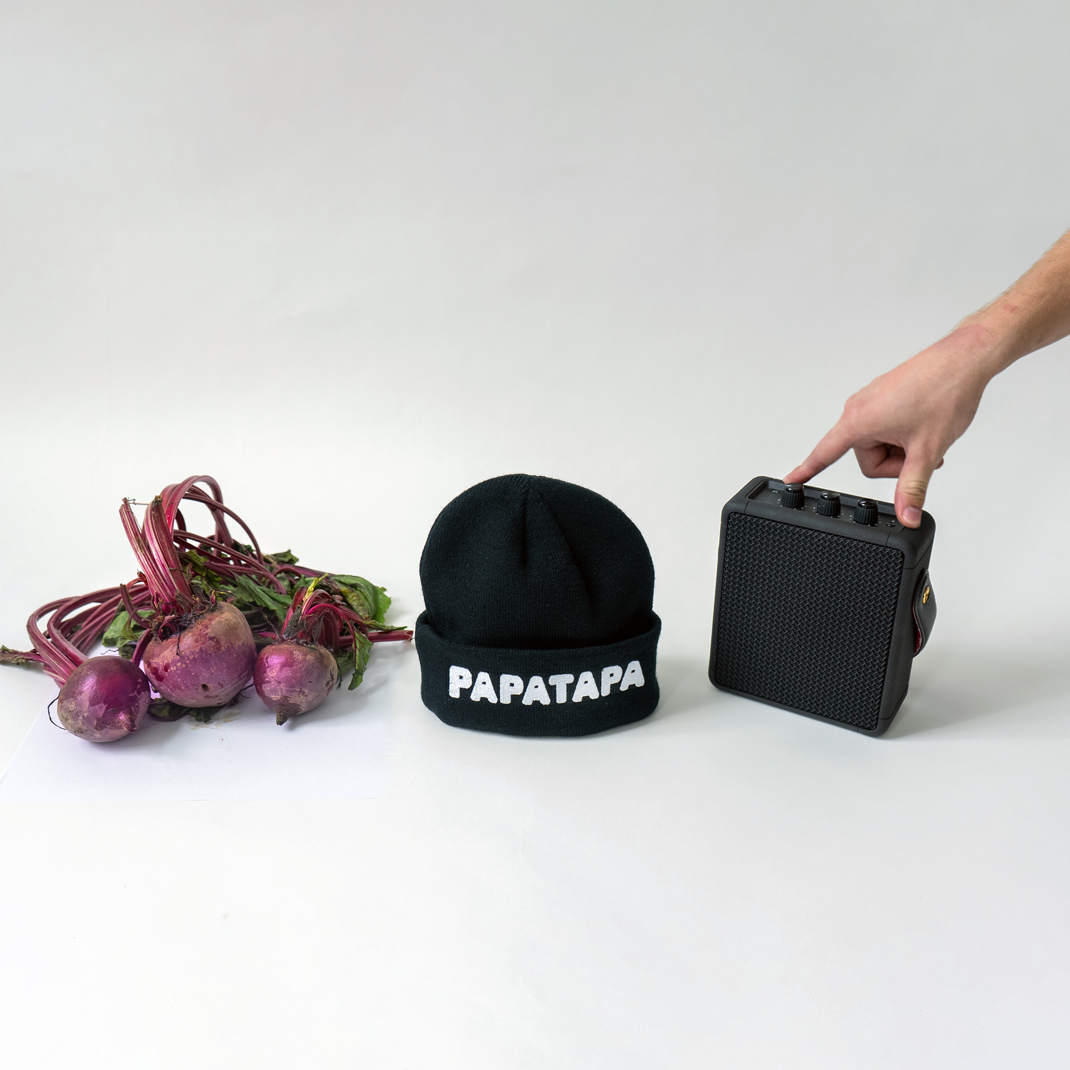 A bunch of beets with leaves, a black beanie with white text 'PAPATAPA', a black portable speaker, and a person's hand adjusting the speaker on a plain white surface.