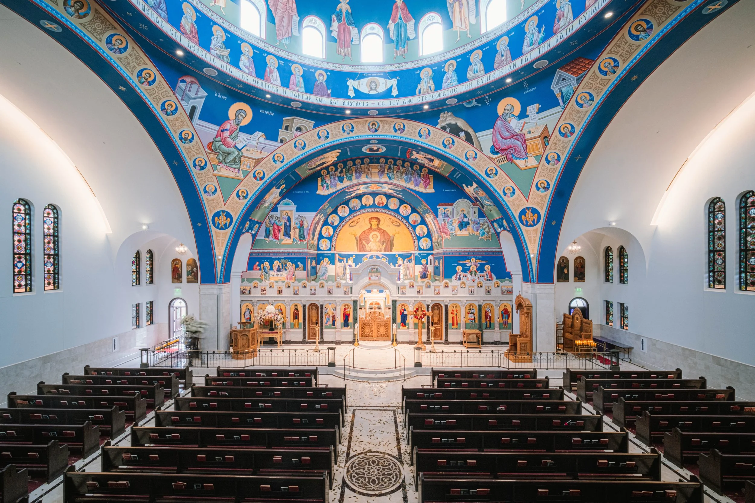 Interior of an Orthodox Christian church with vibrant blue and gold religious murals on the ceiling and walls, stained glass windows, and wooden pews facing an ornate altar area.