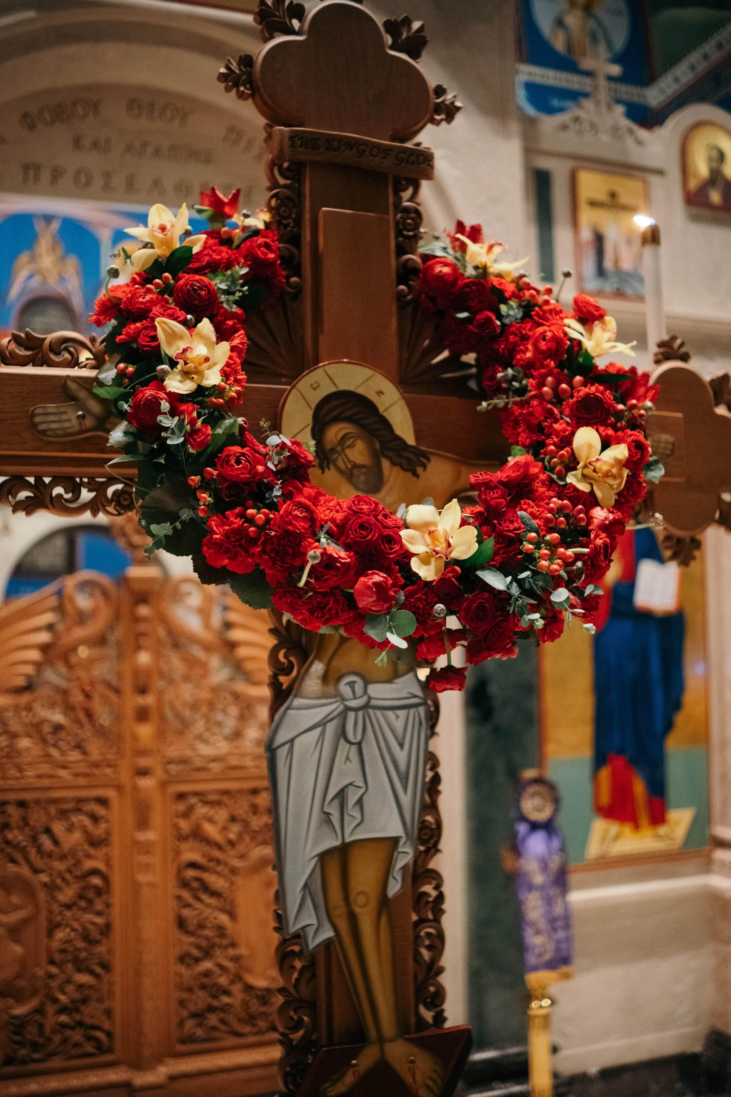 Wooden crucifix with a portrait of Jesus Christ, decorated with a circular wreath of red roses, cream-colored lilies, and greenery inside a church.