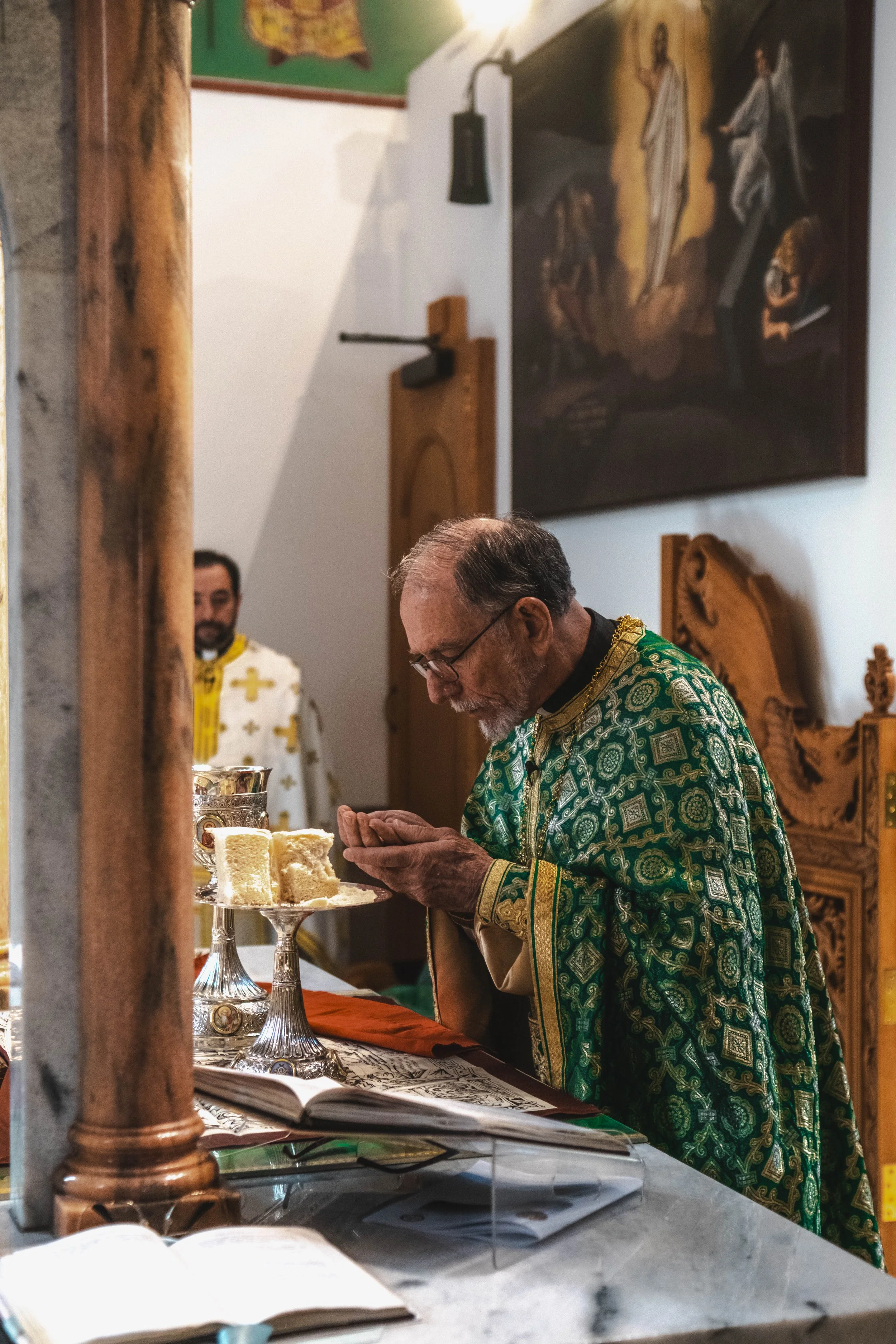 An elderly priest in a green ornate vestment performing a religious ceremony at an altar with bread and candles, with a younger man in a white vestment with gold crosses in the background in a church setting.