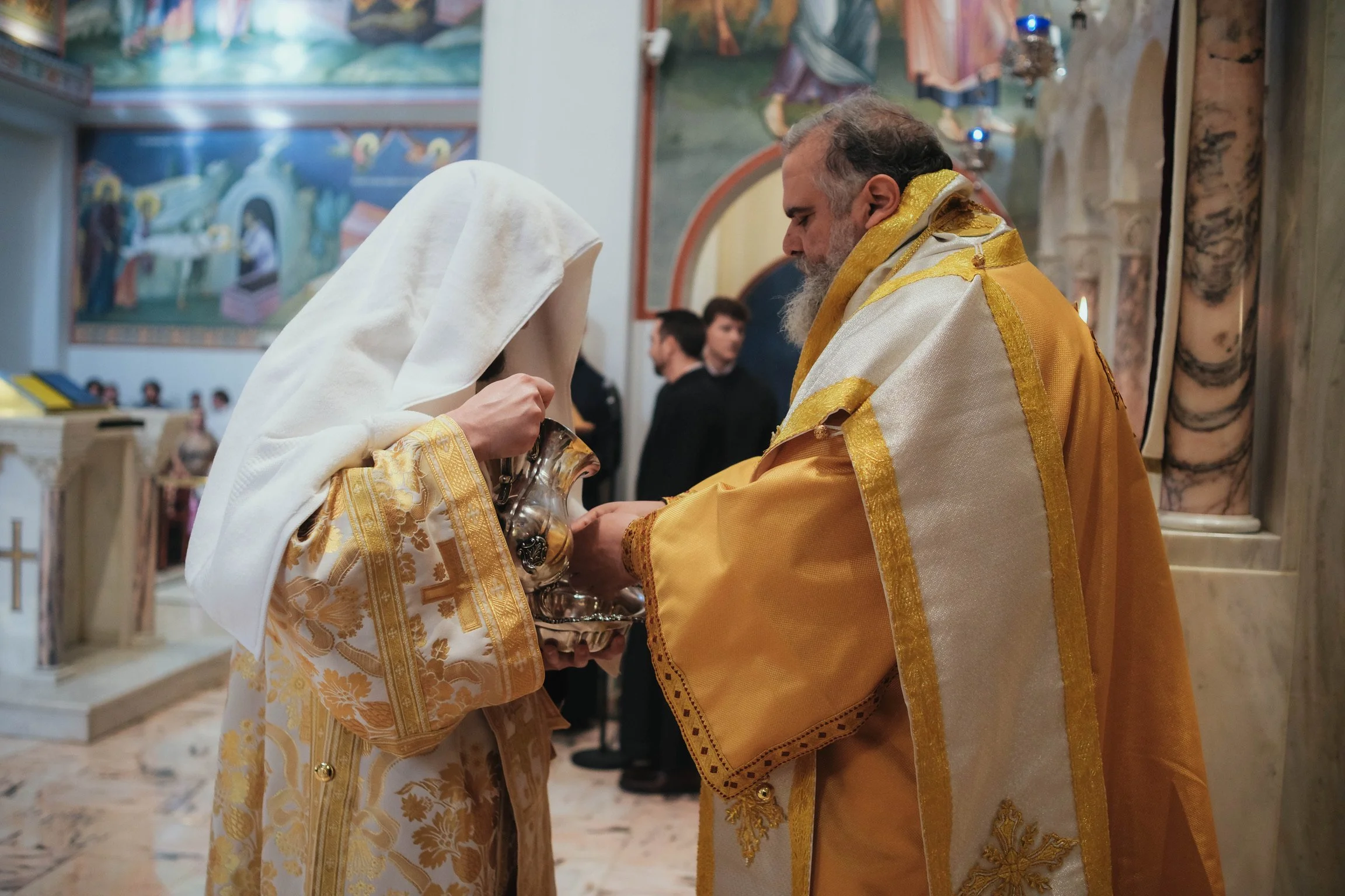 A priest and a woman in ornate religious robes are holding a chalice in a church interior with colorful religious murals.