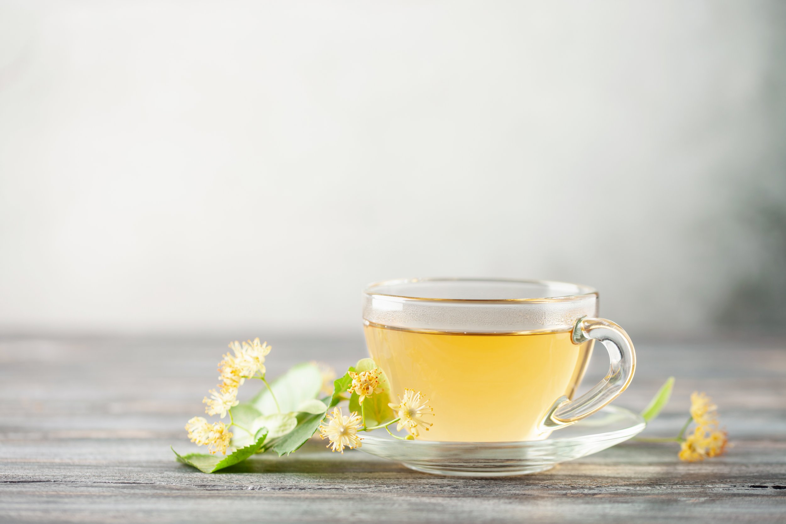 A clear glass cup filled with light green tea on a wooden surface, with small light yellow flowers and green leaves placed beside it.