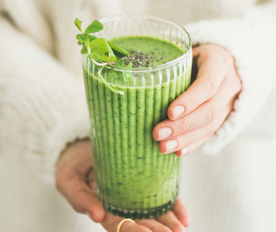A person holding a tall glass of matcha green smoothie garnished with a sprig of fresh herbs, with a creamy white sweater in the background.