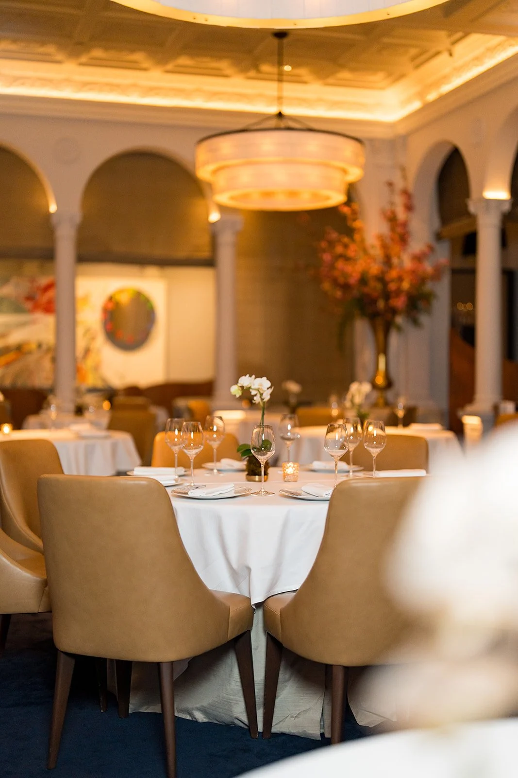 Elegant restaurant interior with round tables set with white tablecloths, wine glasses, and flower centerpieces, illuminated by warm lighting from modern circular pendant lights.