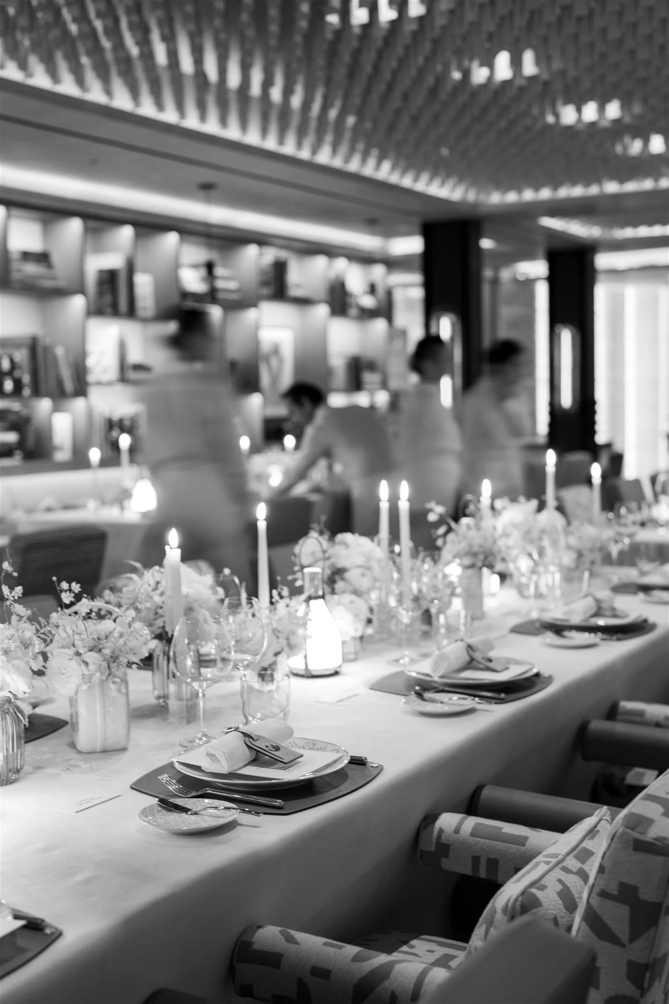 A decorated dining table set with candles, floral centerpieces, wine glasses, and tableware in a restaurant with staff working in the background.