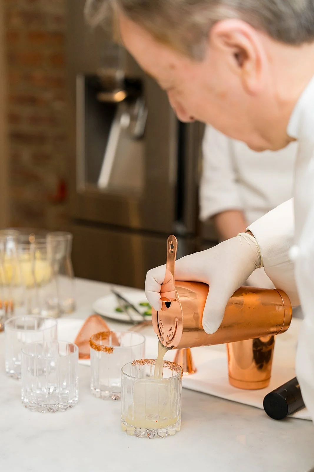 A bartender in white gloves pouring a cocktail from a copper shaker into a glass with a salted rim.