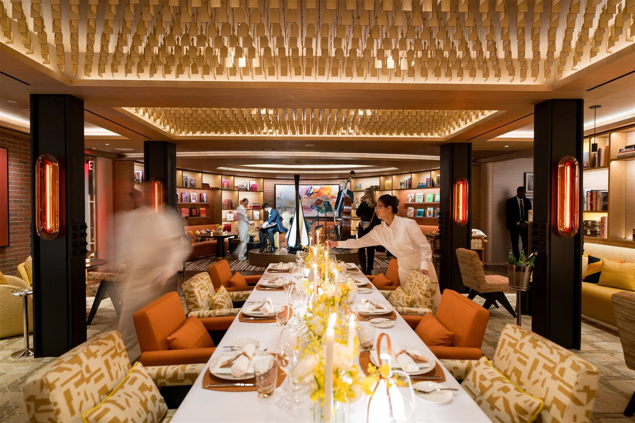 Elegant dining room with a long table set for a meal, surrounded by modern patterned chairs. The room features warm lighting, a bookshelf with books, and decorative wall elements. Several people, including staff, are preparing the space for a gatheri