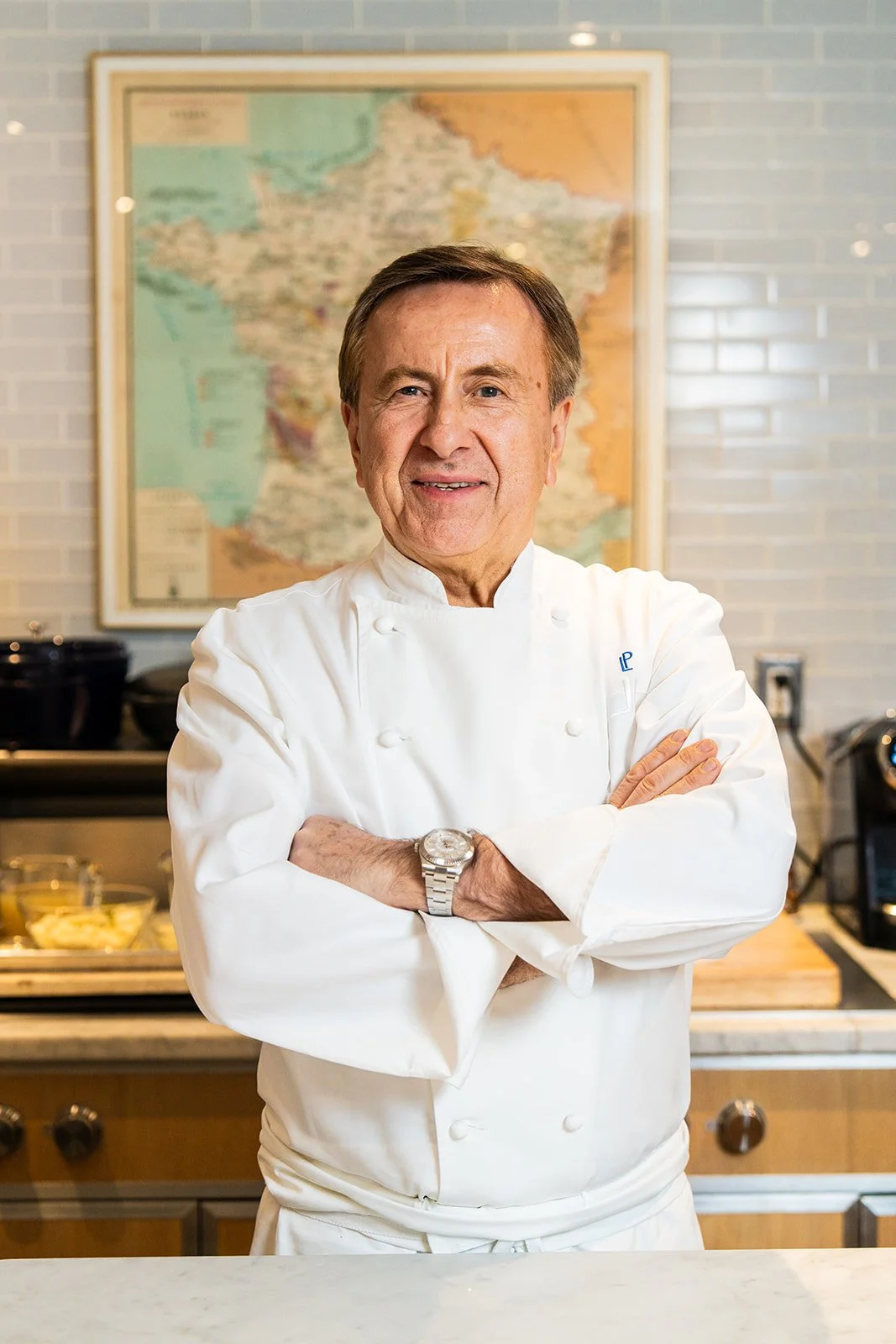 A smiling man in a white chef's coat with crossed arms in a kitchen with a map of France on the wall behind him.